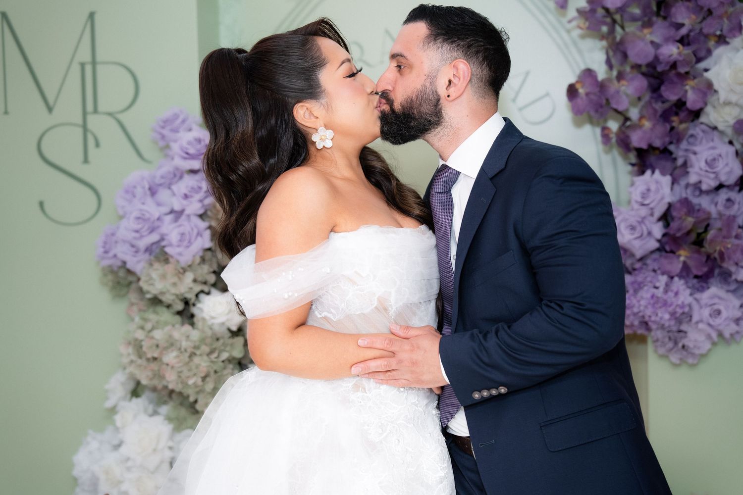 Bride and groom sharing a kiss in front of a floral wall backdrop with purple flowers at their wedding.