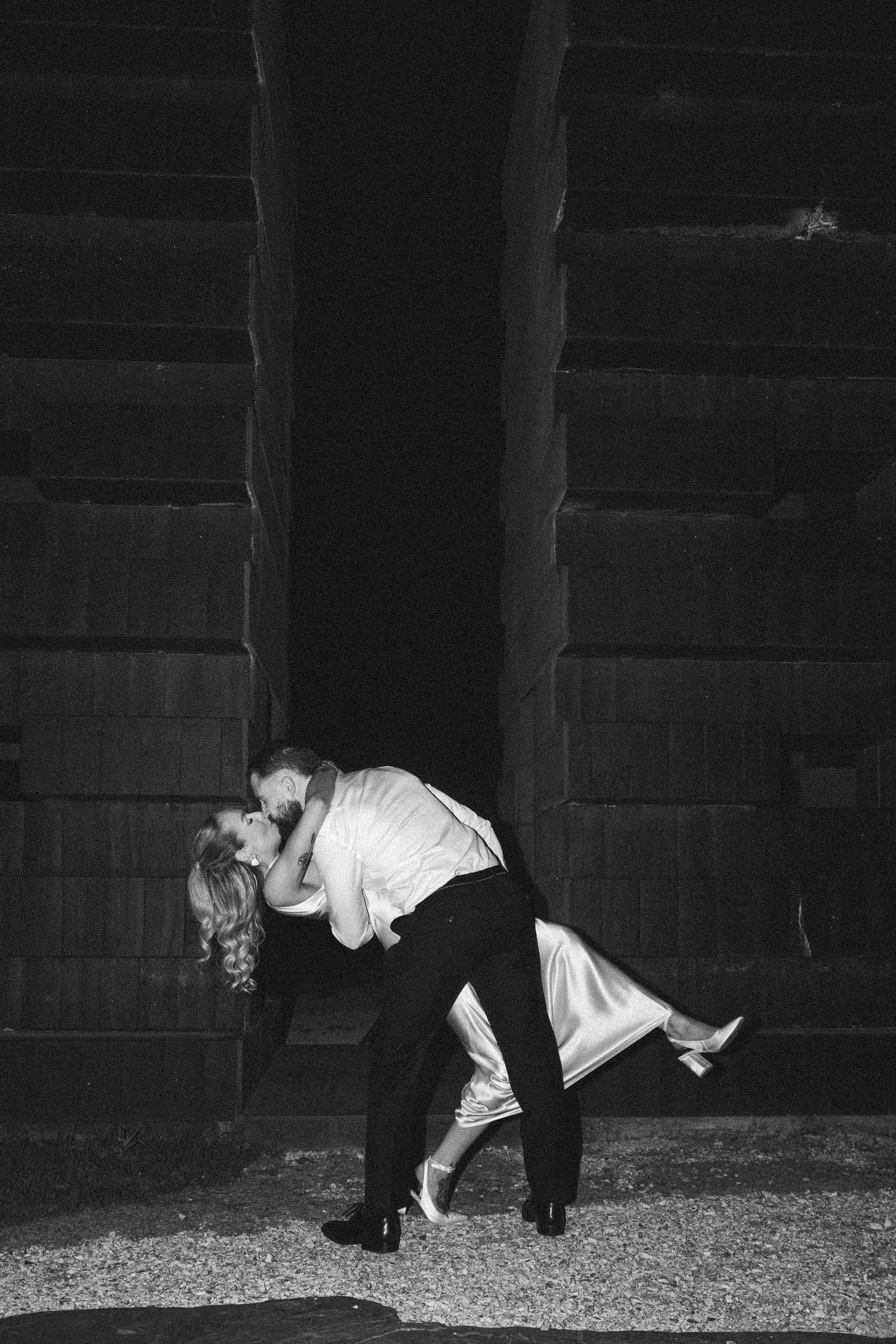 A romantic dip pose captured in black and white against an industrial backdrop.