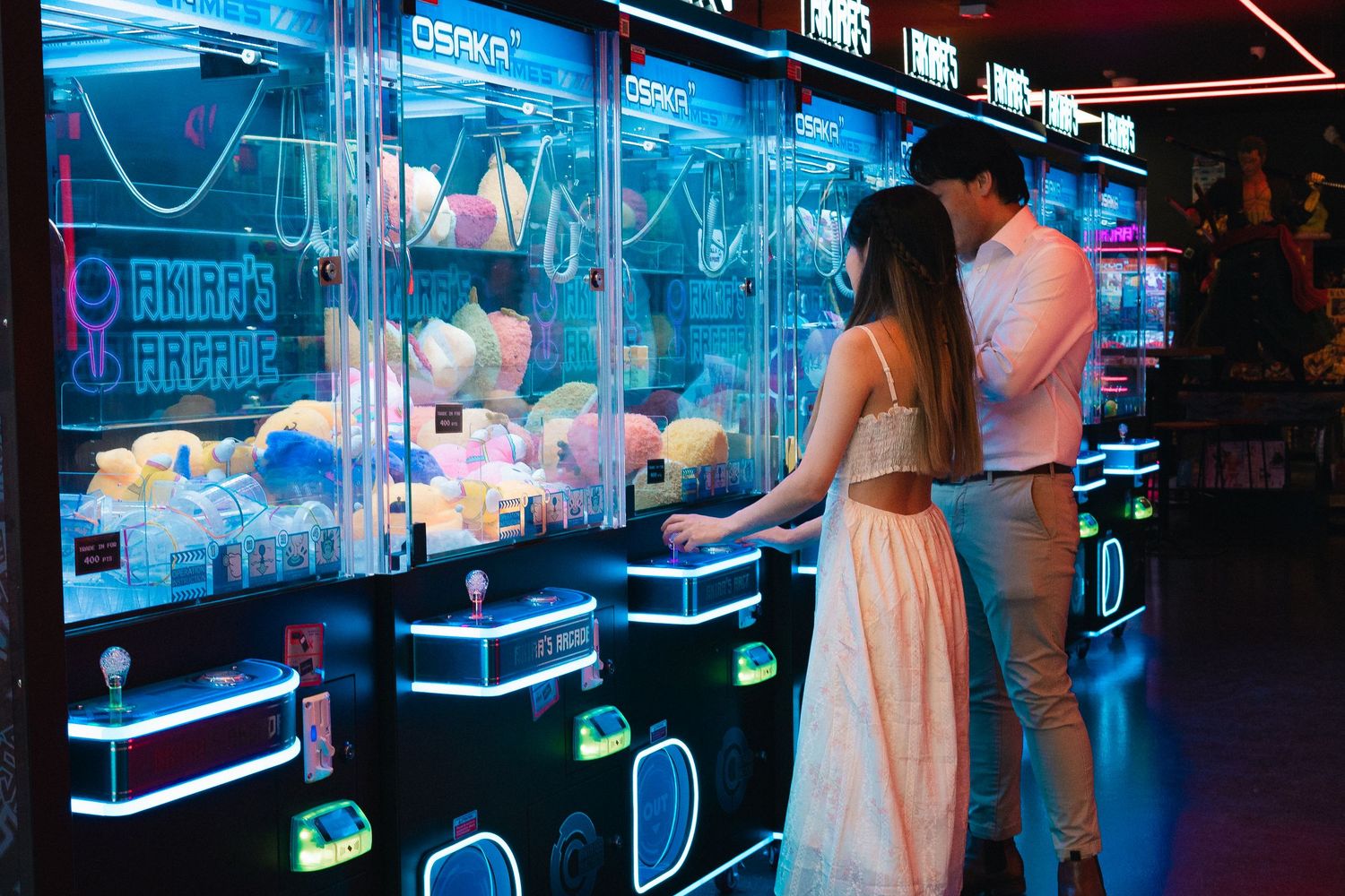 Couple playing claw machine games at a neon-lit arcade filled with colorful stuffed animal prizes.