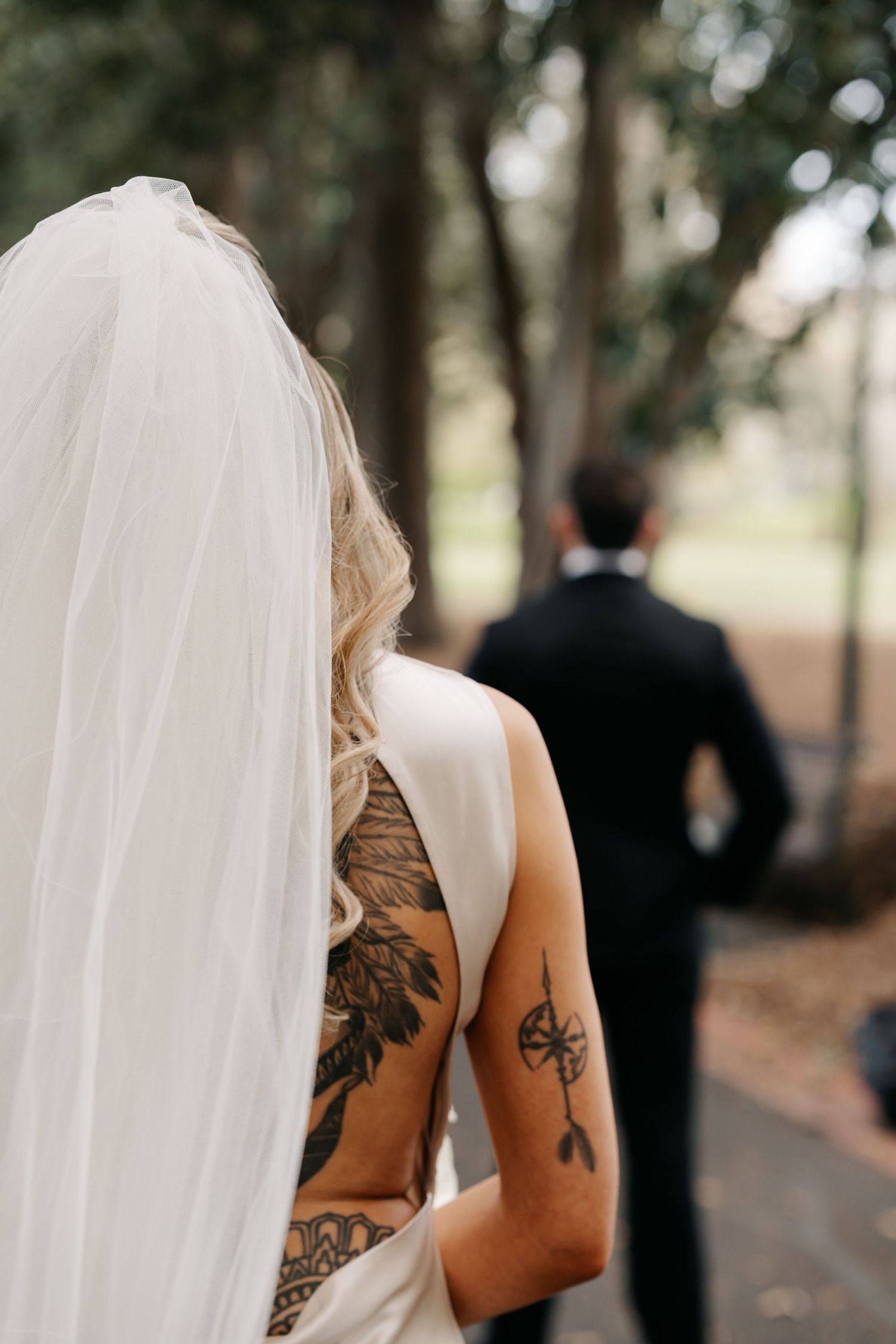 Bride with tattoos and flowing veil walking away from groom in outdoor setting.