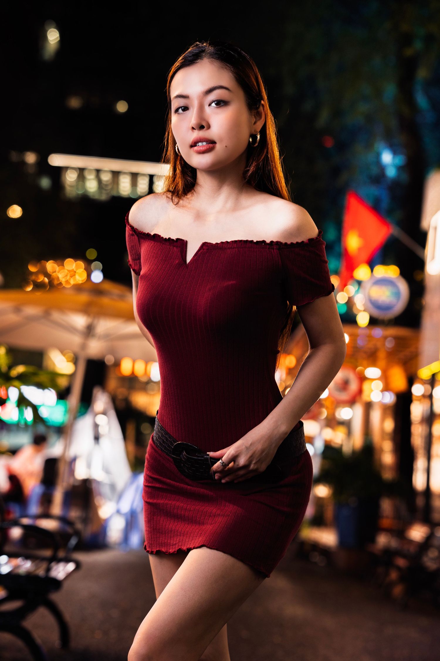 Woman in burgundy dress poses dramatically against city lights at night.