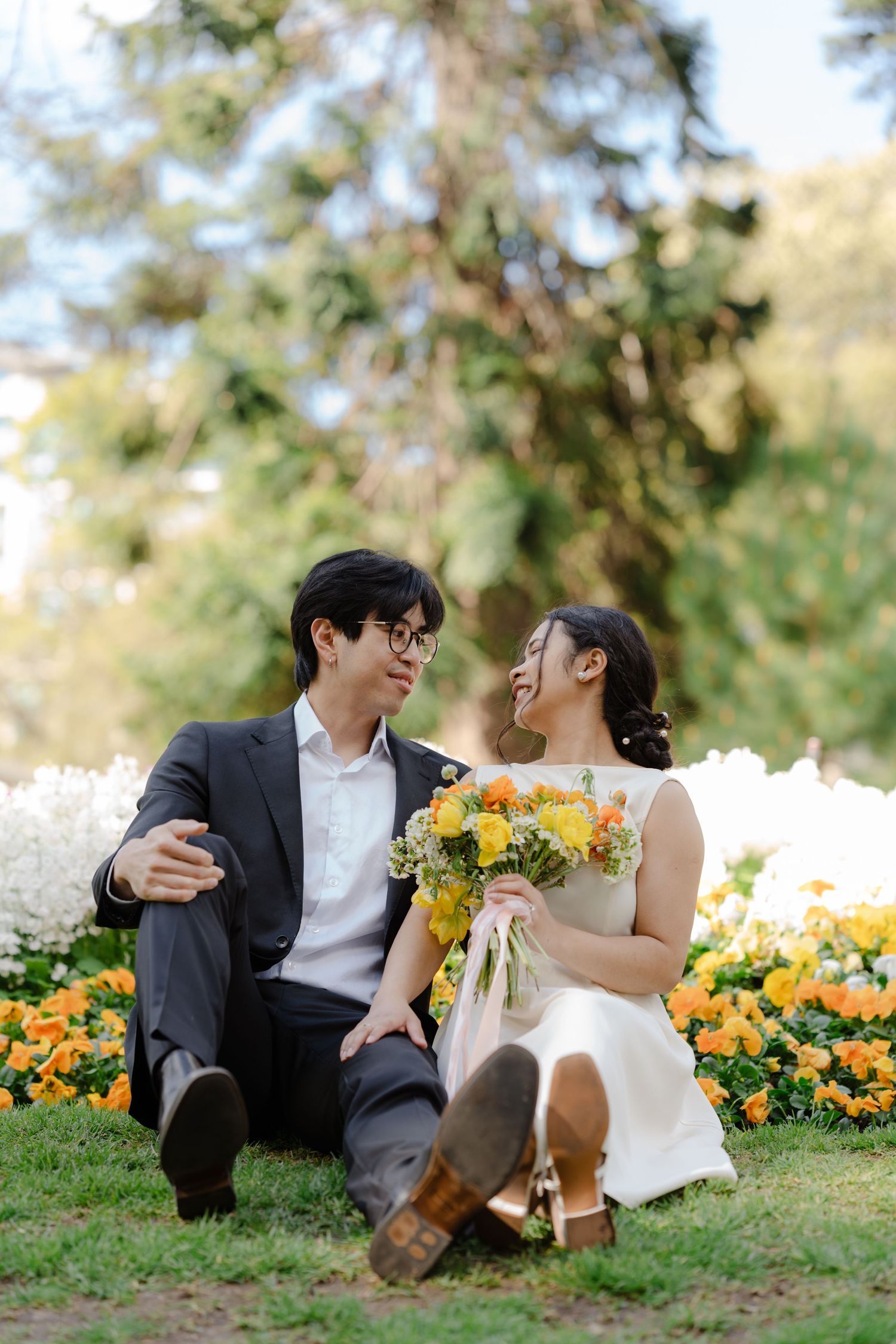 A couple sits on grass surrounded by orange and yellow flowers in a garden setting.