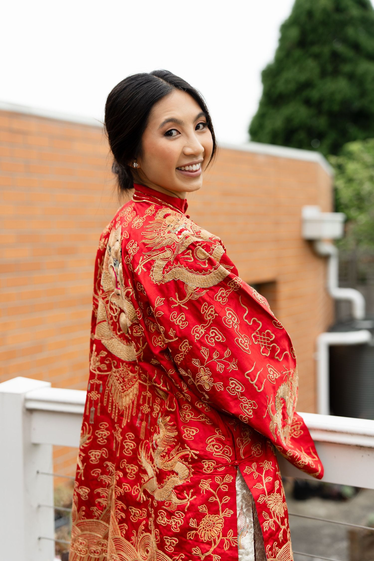 Smiling Asian woman in red and gold embroidered traditional Chinese qipao dress posing outdoors on a balcony.
