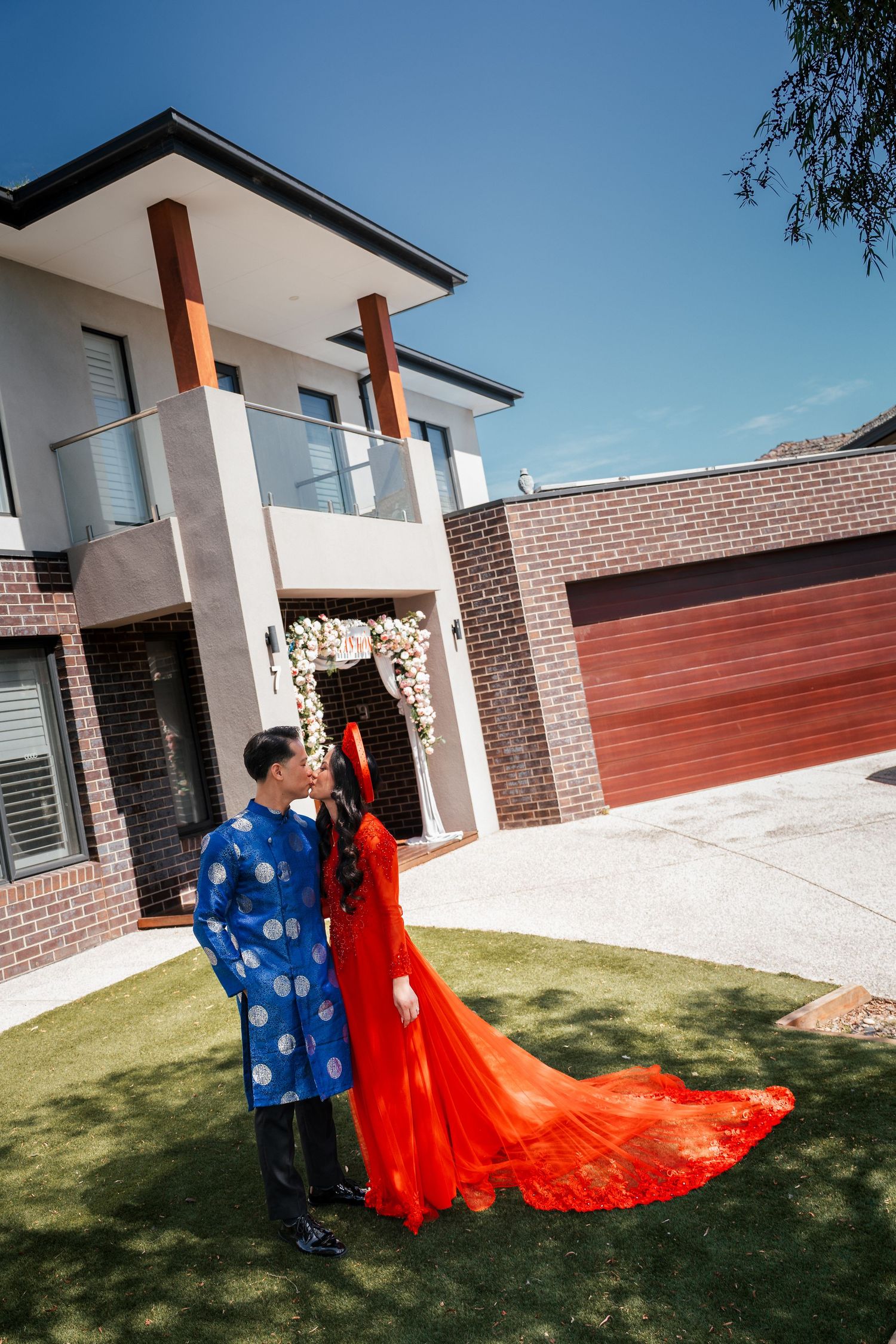 Couple in traditional Vietnamese ao dai attire posing outside a modern brick home on a sunny day.