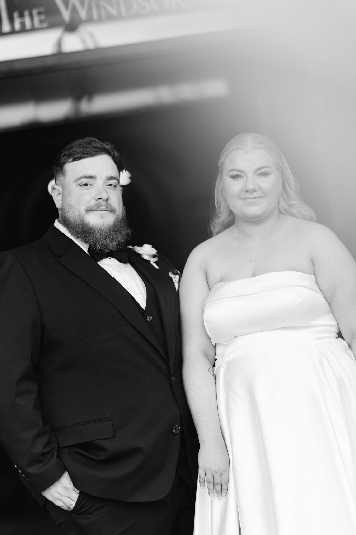 Bearded groom in tuxedo and bride in strapless gown pose outside The Windsor in black and white wedding portrait.