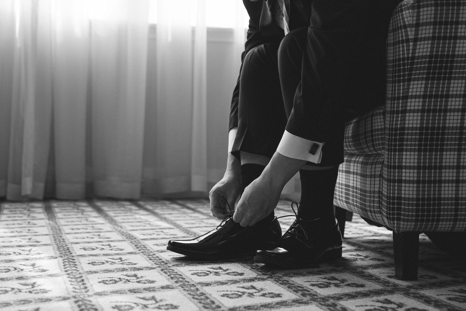 Black and white photo of a man tying his dress shoes, wearing a suit with cufflinks, seated near a curtained window.