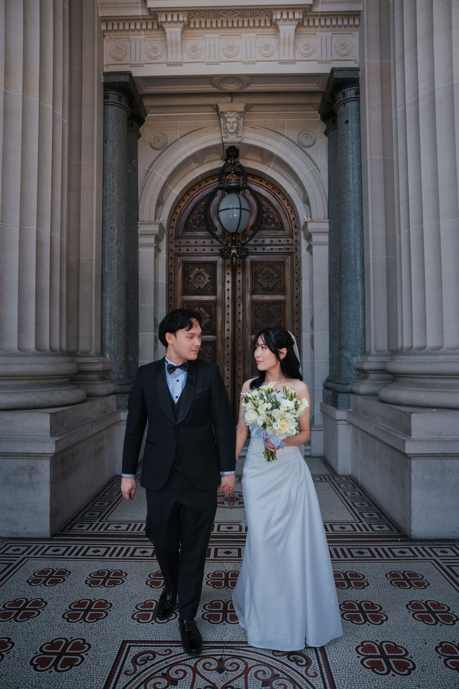 Elegant couple in wedding attire posing before ornate bronze doors and grand columns on decorative tile floor.
