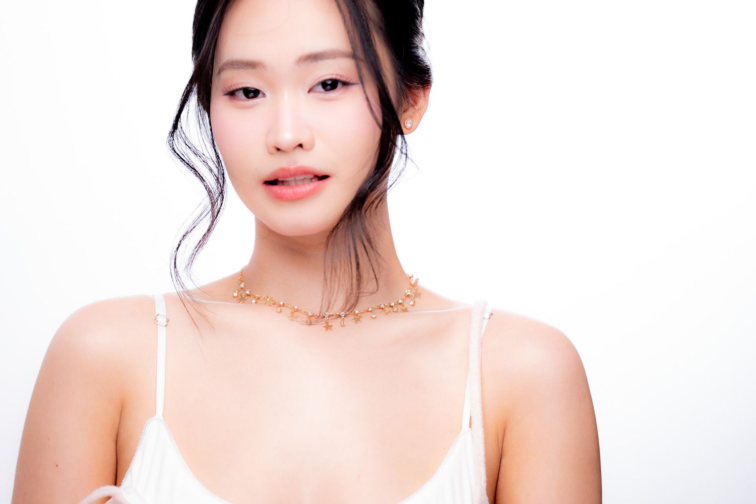 Young Asian woman in white camisole wearing gold necklace, posing against white background.