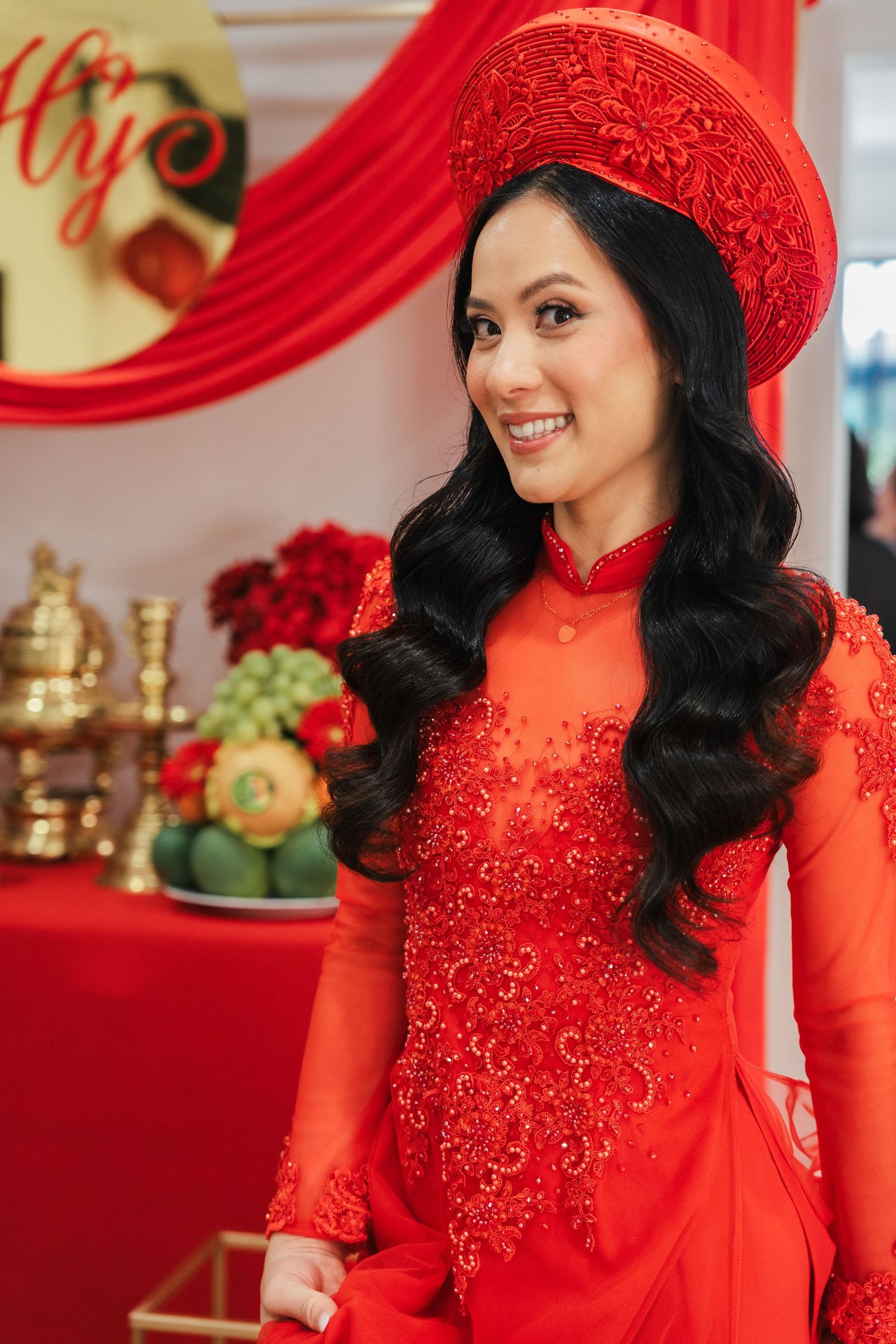 Vietnamese bride smiling in red ao dai dress and non la hat at traditional wedding ceremony.
