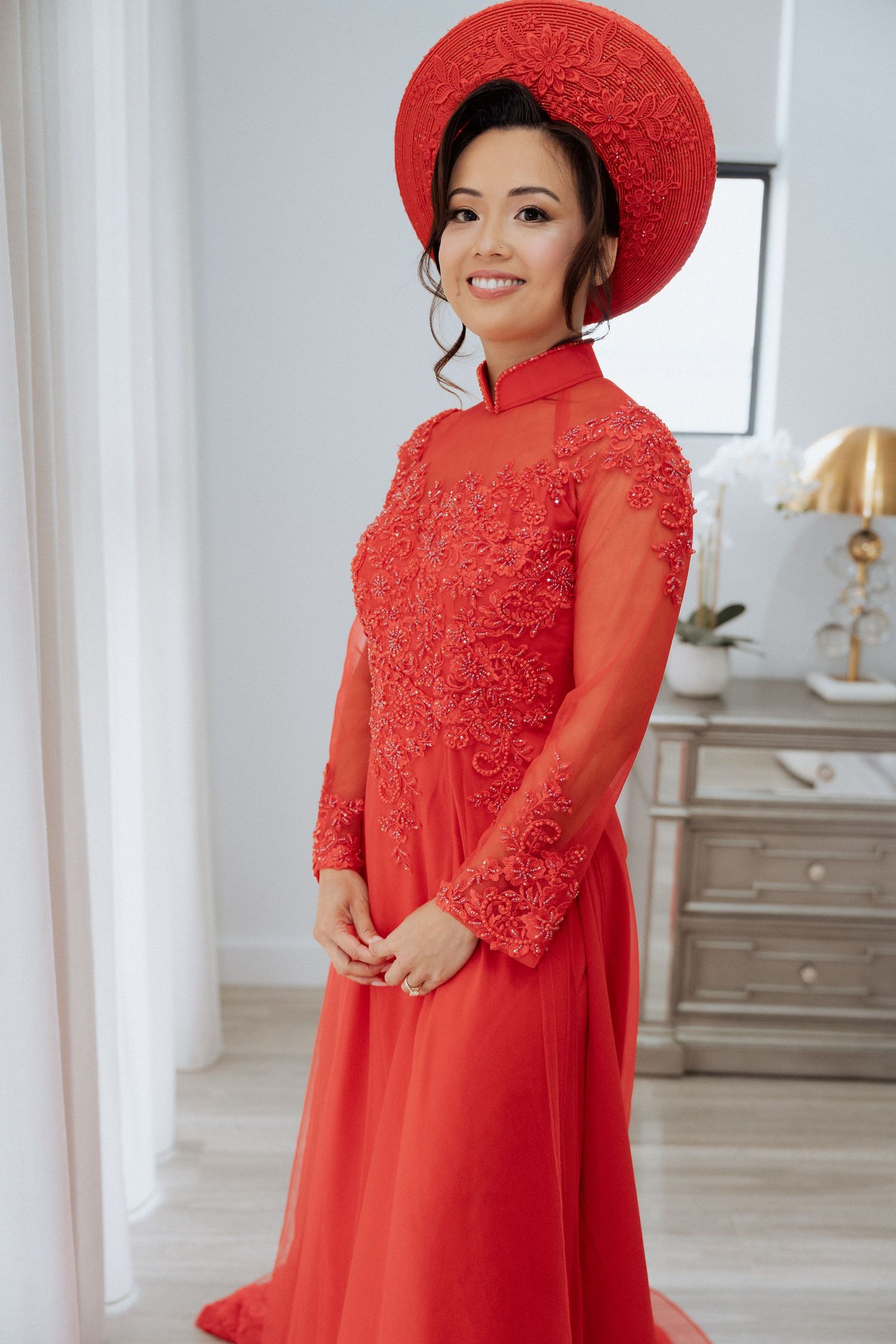 Elegant woman posing in a red Vietnamese Ao Dai dress with floral lace bodice and traditional red conical hat.