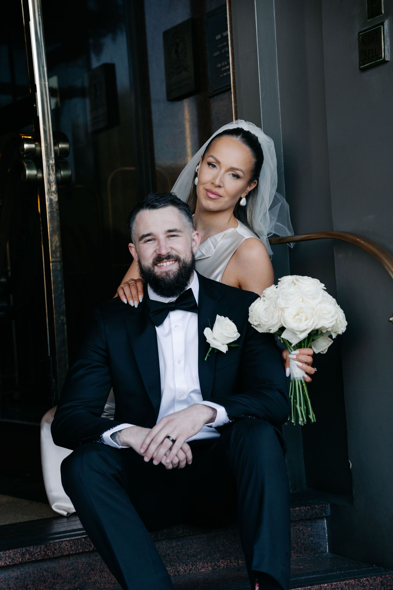 Wedding couple poses together on steps with white roses and formal attire.