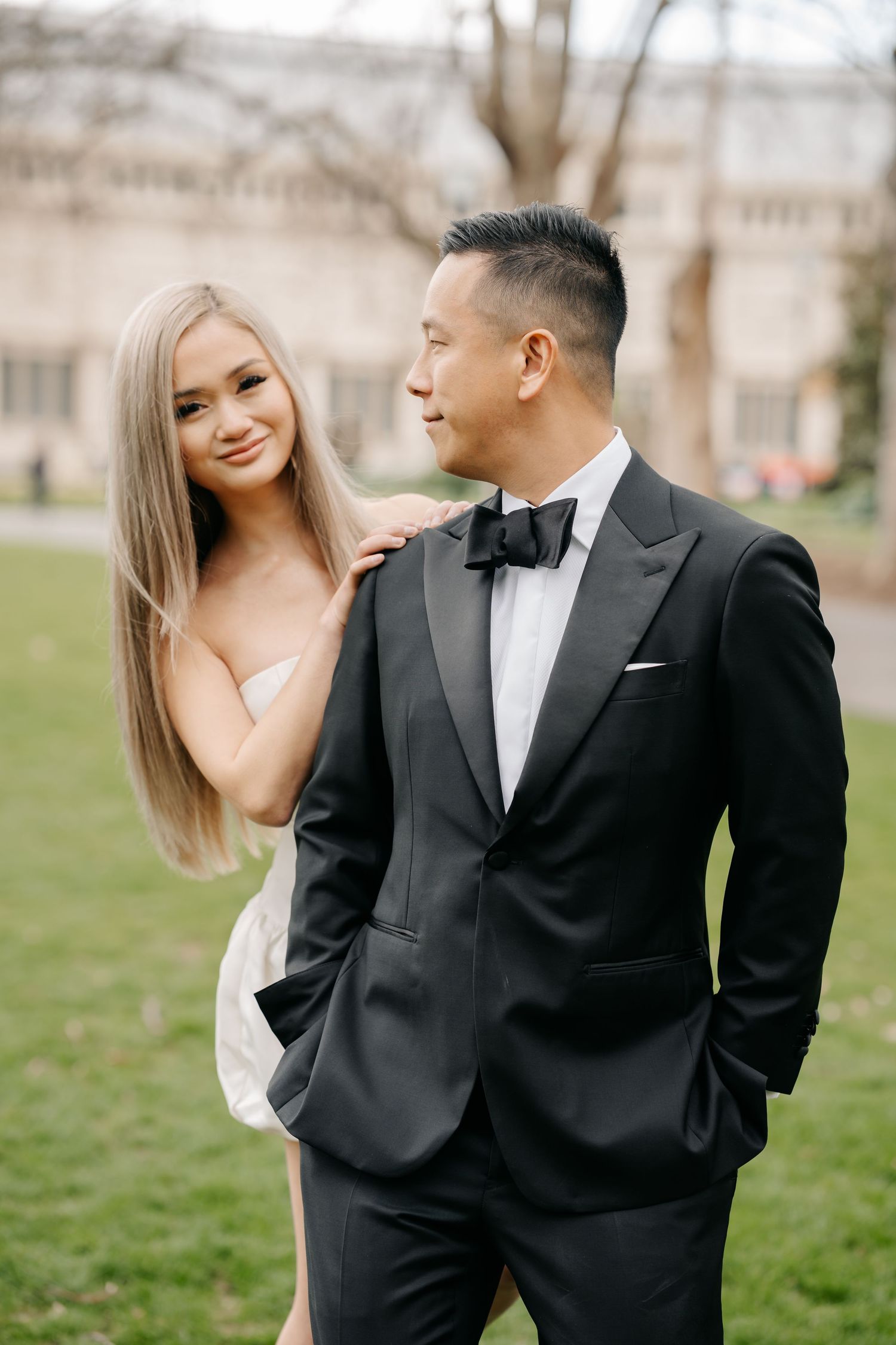 Couple in formal attire shares intimate moments on a grassy lawn with a grand building in the background.