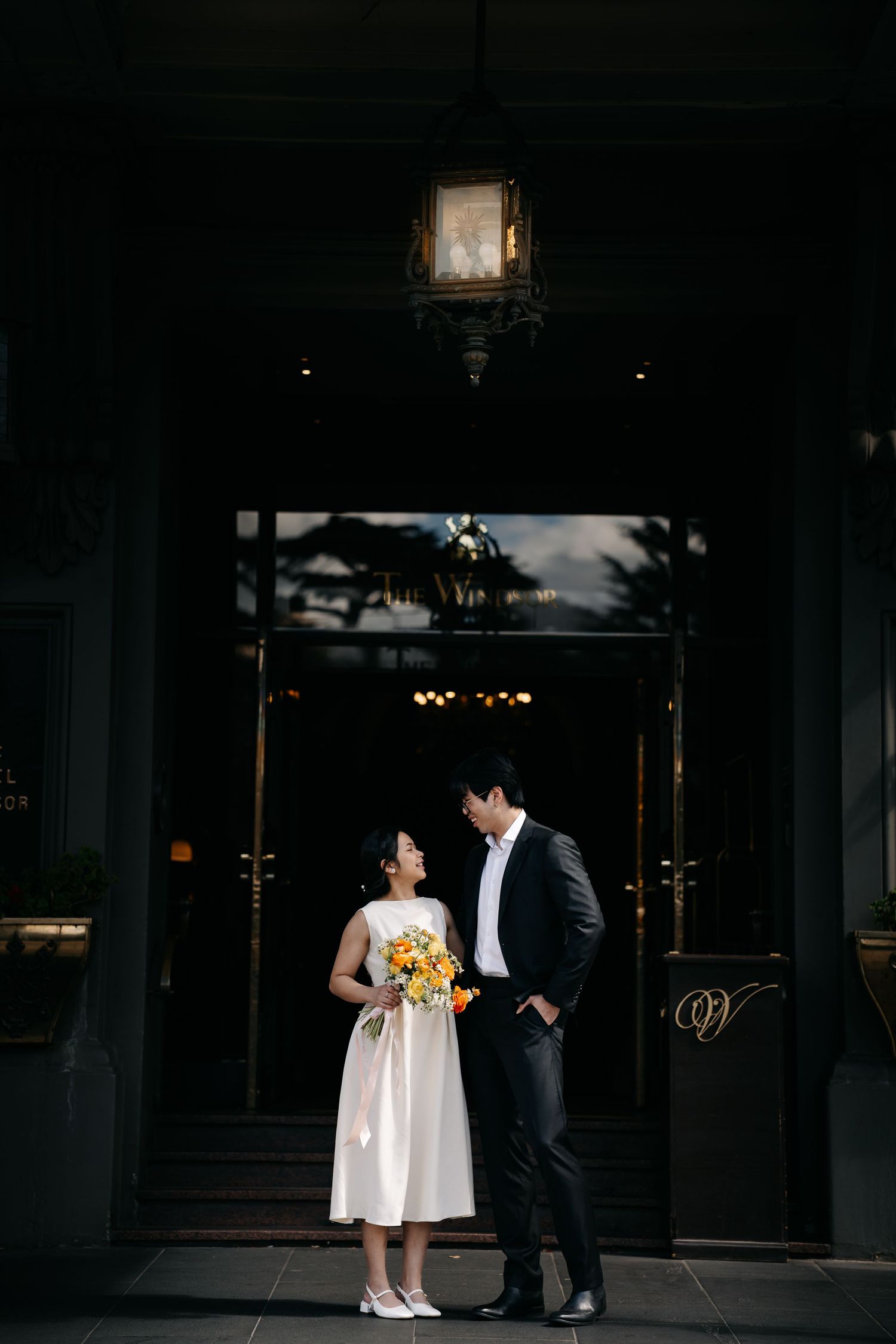 A couple in wedding attire stands in dramatic shadows against a dark background with soft lighting.