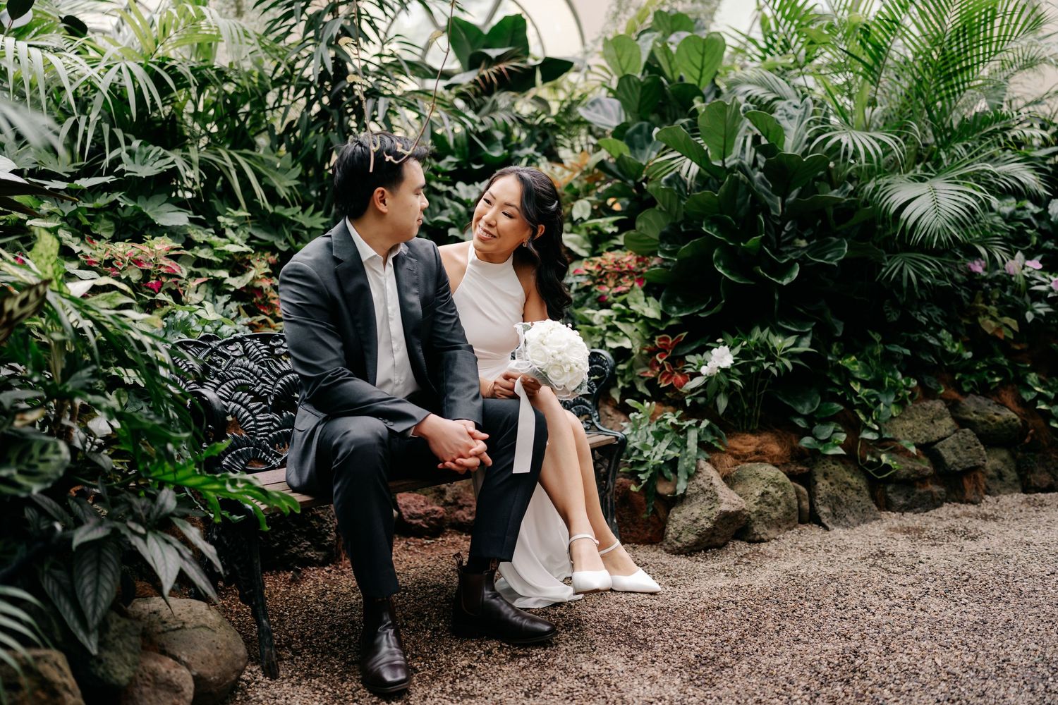 A couple in wedding attire sits on a bench surrounded by lush tropical greenery, sharing a tender moment.