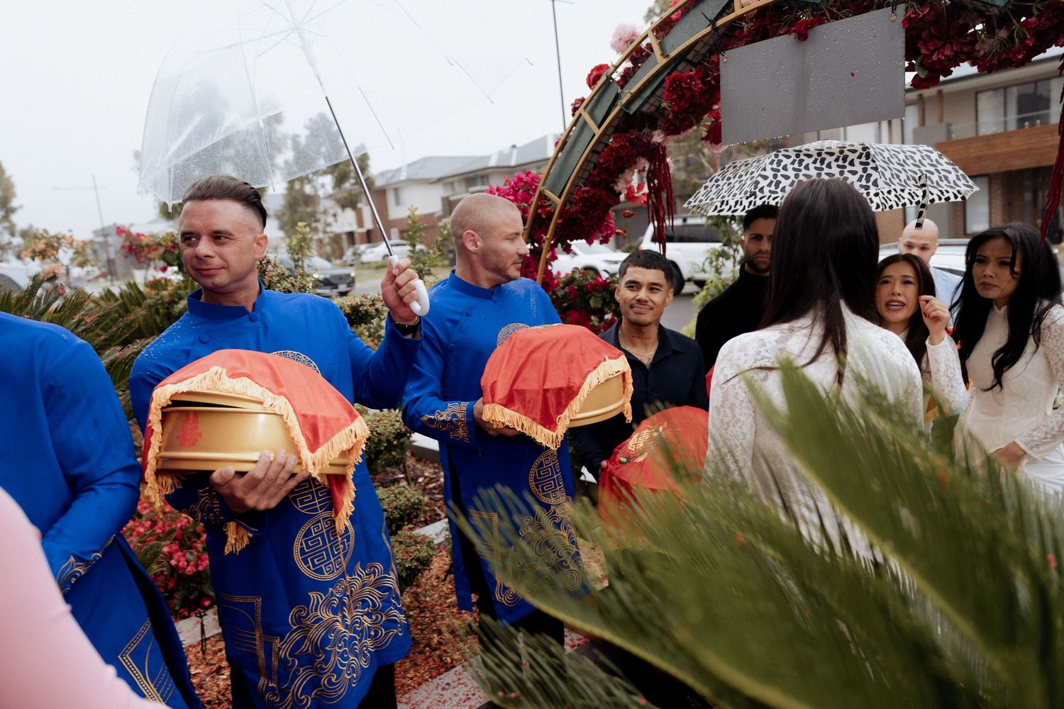 Men in blue ao dai carry red ceremonial boxes in a festive outdoor procession with floral decorations and umbrellas.