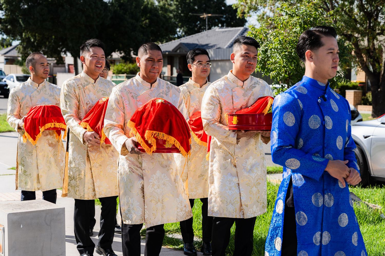 Vietnamese groomsmen in gold ao dai carrying red gift trays at traditional wedding ceremony outdoors.
