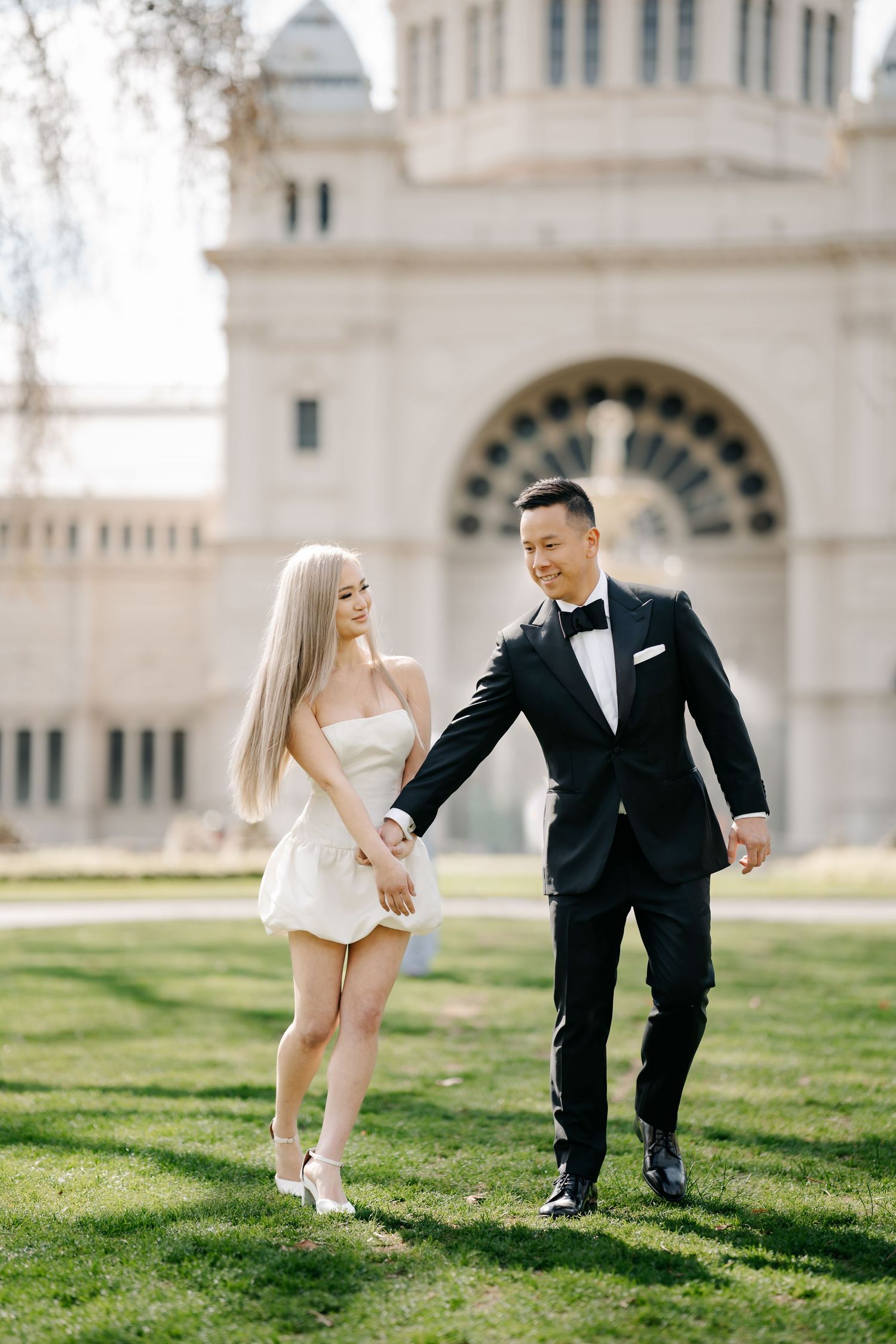 Couple playfully interacts on a sunny lawn with historic architecture visible behind them.