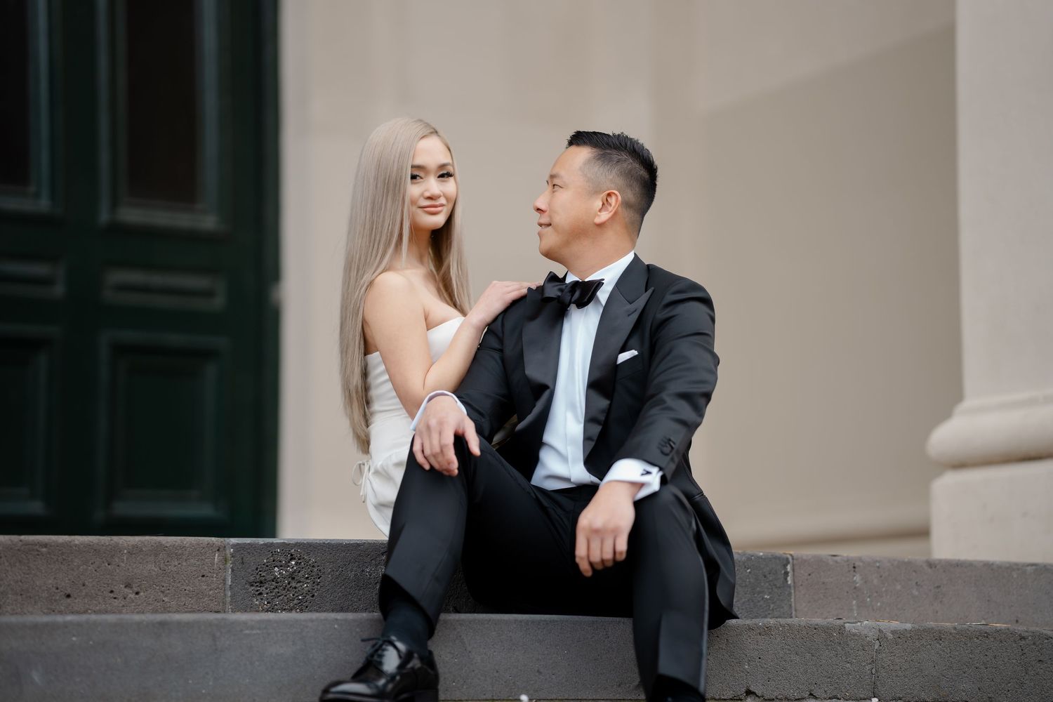 A couple in formal wear sits together on stone steps during their wedding photoshoot.