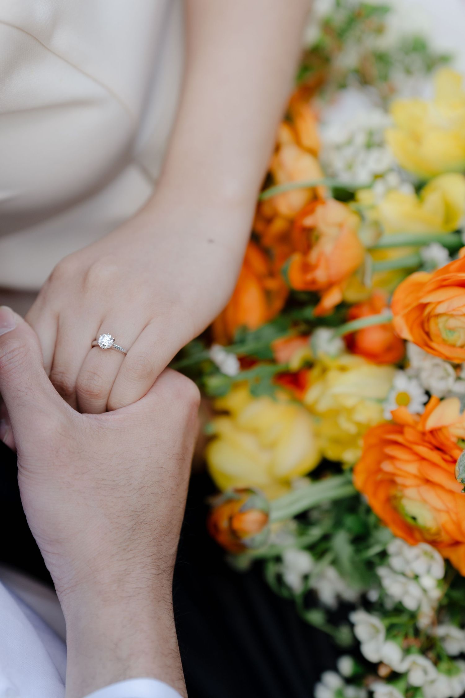 A close-up of hands holding with an engagement ring and colorful floral bouquet in view.