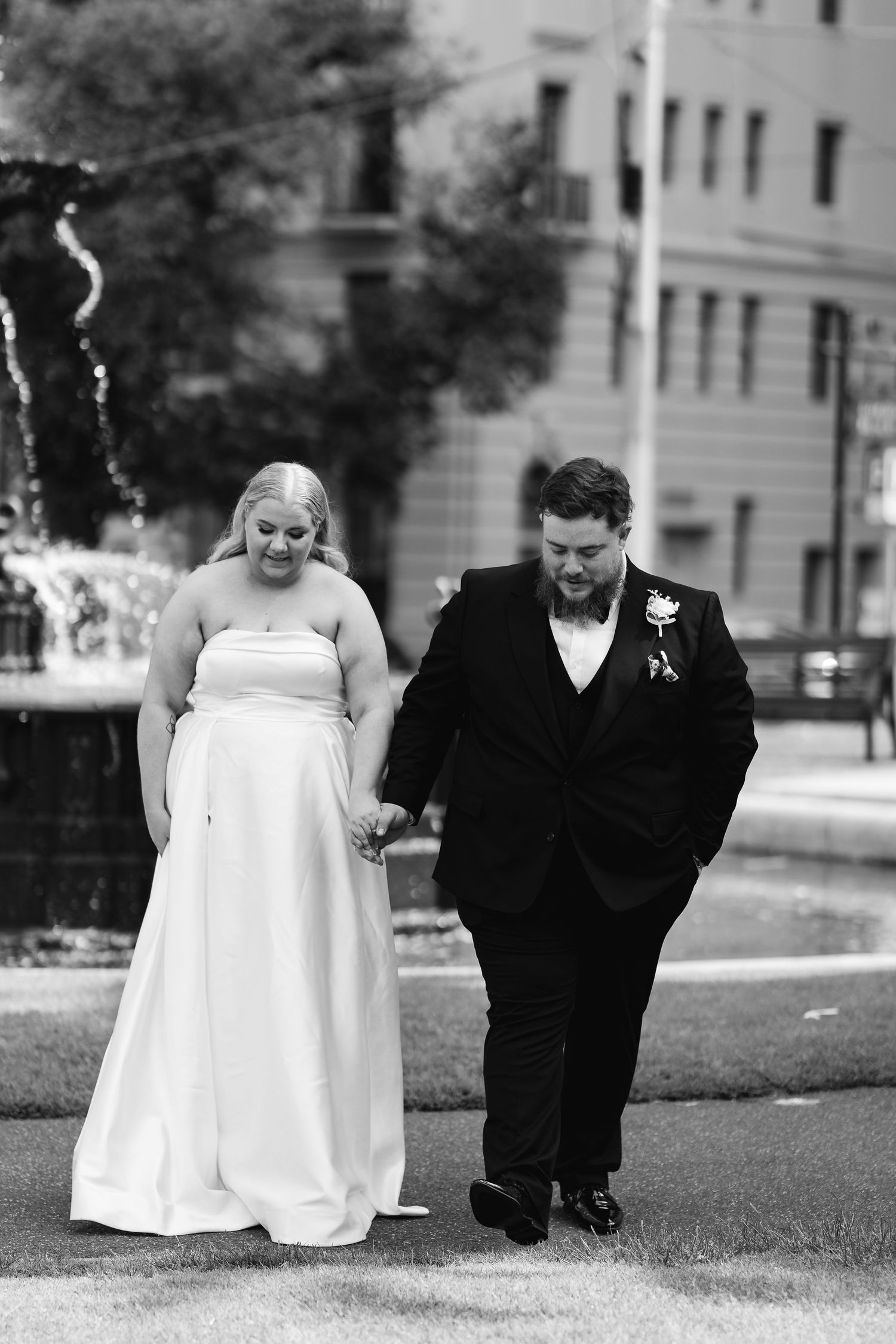 Black and white photo of a bride and groom holding hands walking near a fountain on their wedding day.