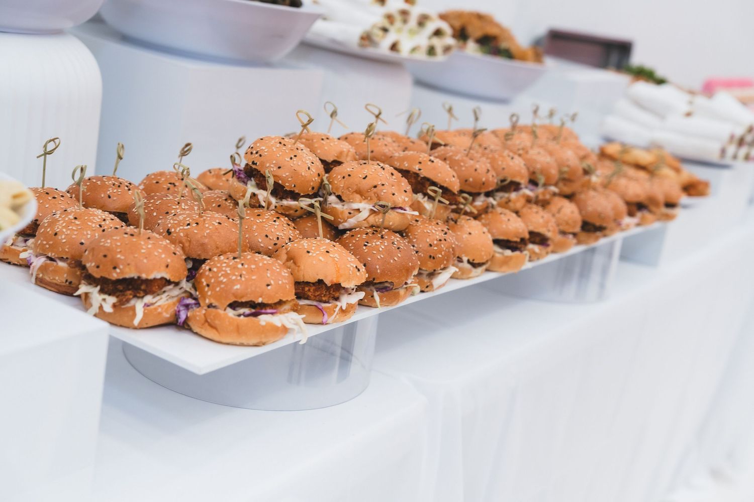 Rows of mini sesame seed bun sliders served on white platters at a catered event.