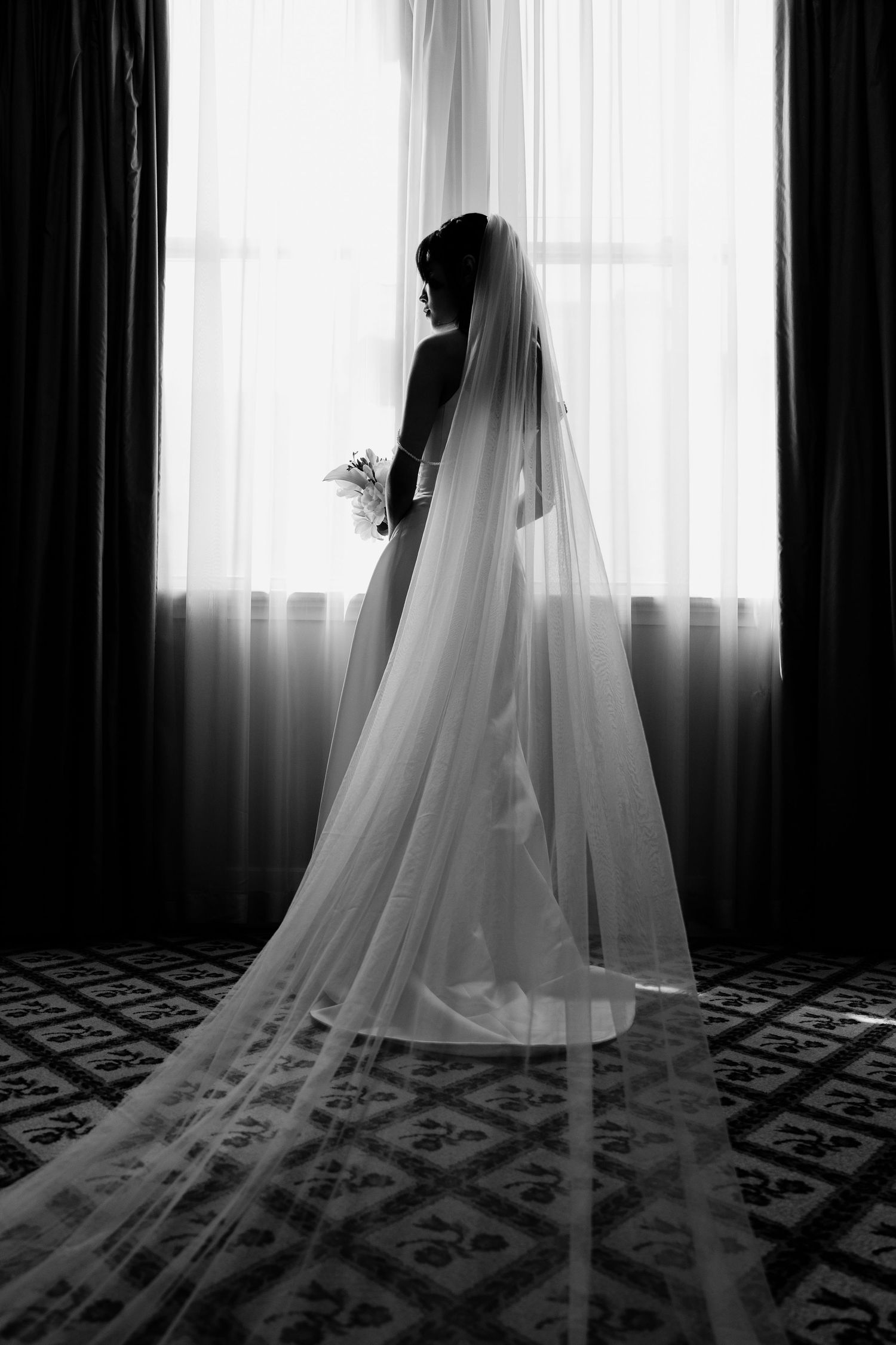 Black and white silhouette of a bride in a long veil standing by a bright window holding a bouquet.