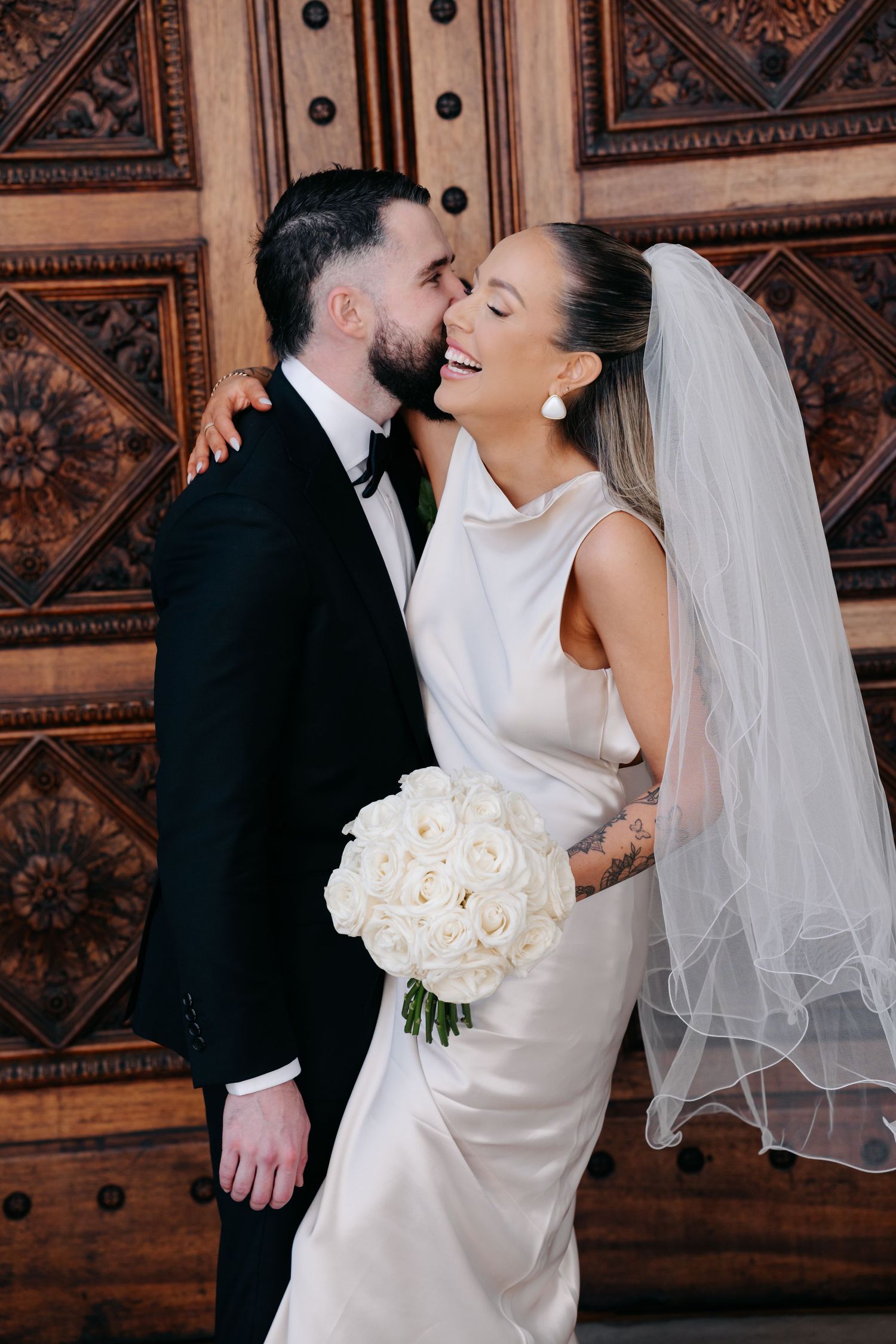 A romantic wedding moment showing a couple embracing in front of an ornate wooden door with the bride holding white roses.