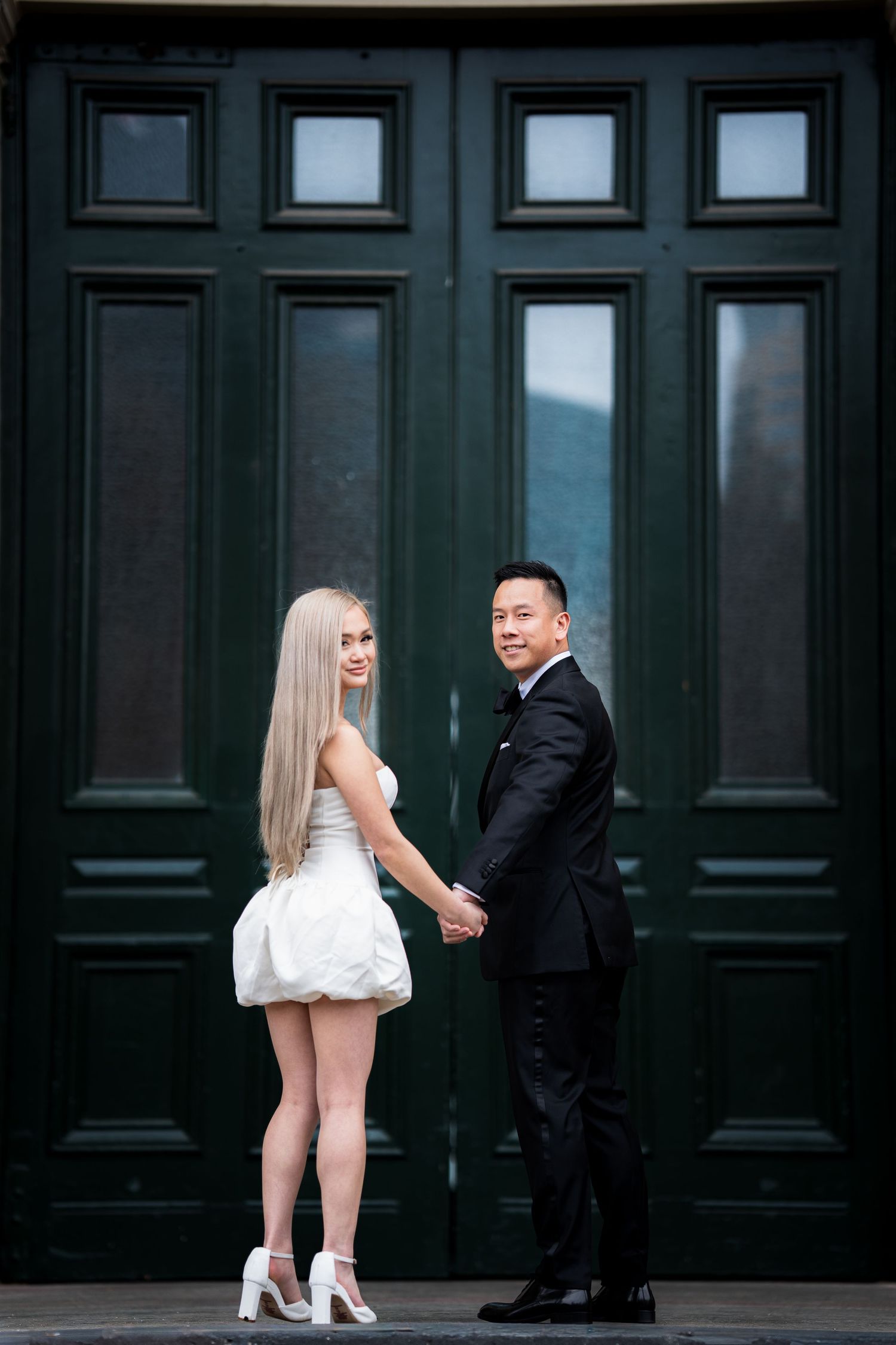 Couple in formal wear poses playfully against dark wooden doors.