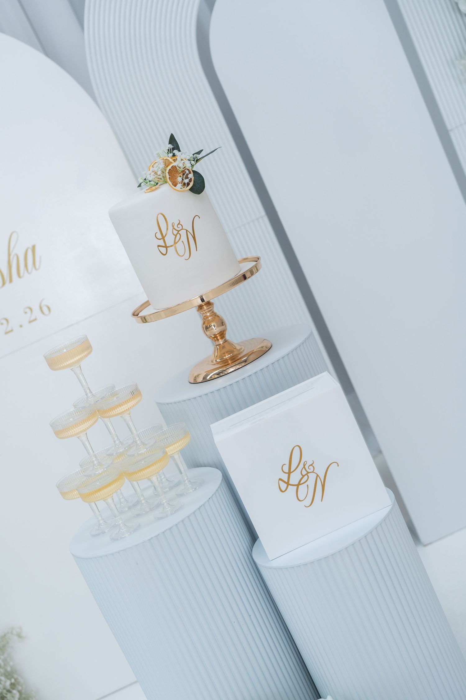 Elegant white wedding cake with gold monogram on a gold stand, surrounded by champagne tower and matching gift boxes.