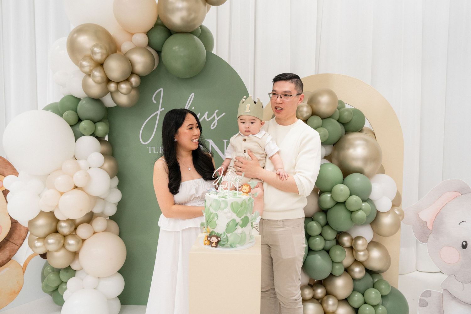 Family celebrating baby's first birthday with sage green and gold balloon arch and decorated cake.