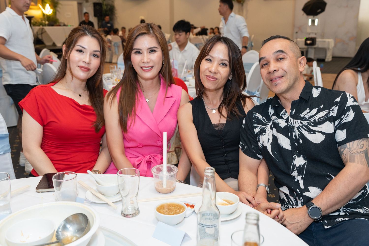 Four friends smiling at a formal dinner event, two women in red and pink dresses, one in black, and a man in floral shirt.