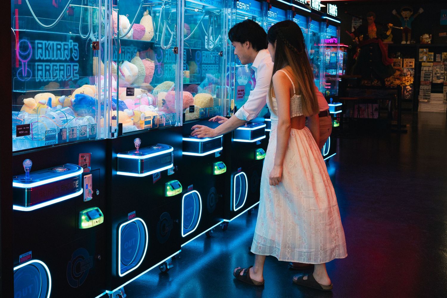 Couple playing claw machine games at a neon-lit arcade filled with stuffed animal prizes.