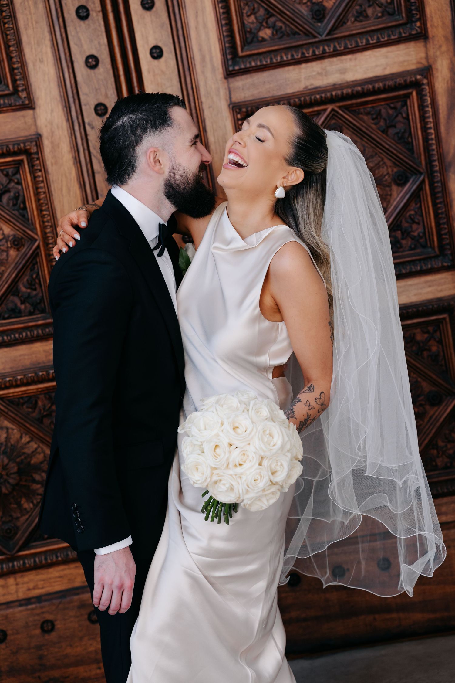 Joyful wedding moment against ornate wooden doors with bride holding white rose bouquet.