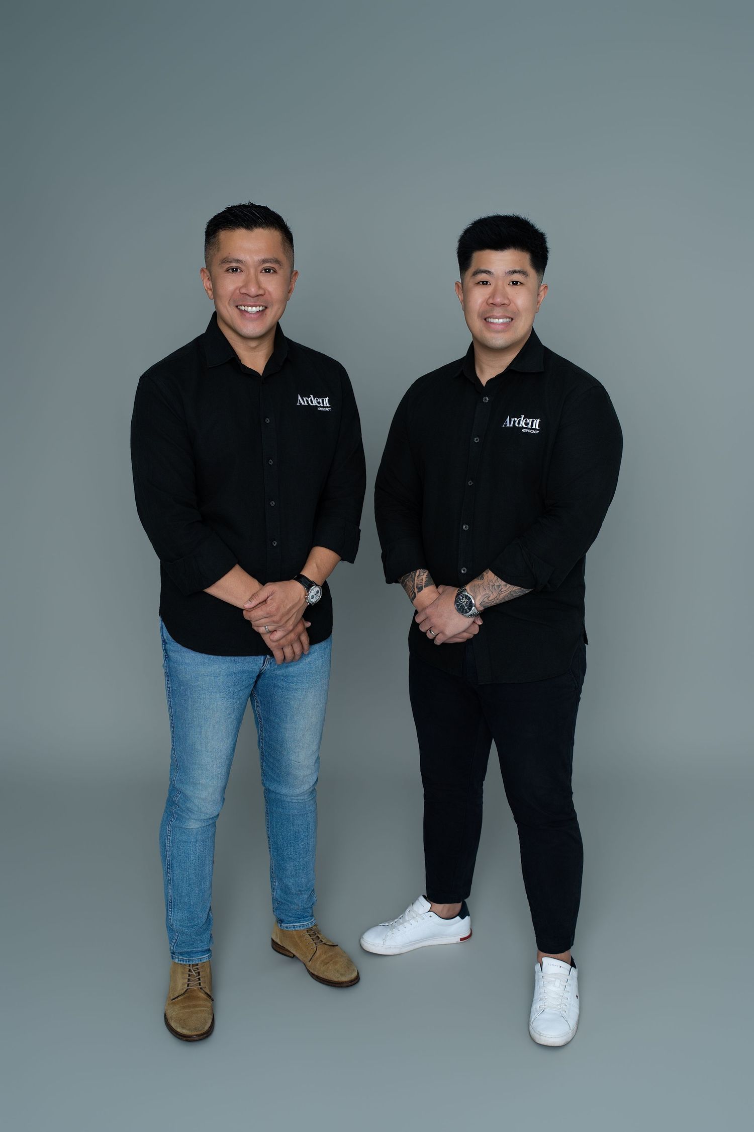Two men in matching black branded shirts pose together smiling against a gray studio background.