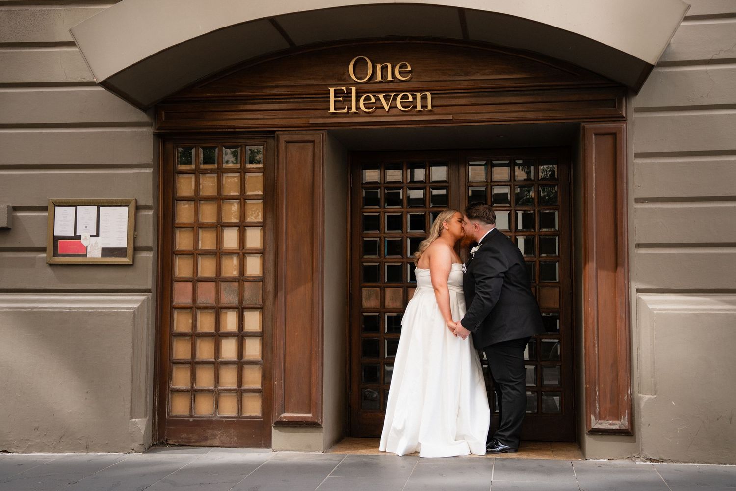 A bride and groom share a kiss in front of the One Eleven venue entrance on their wedding day.