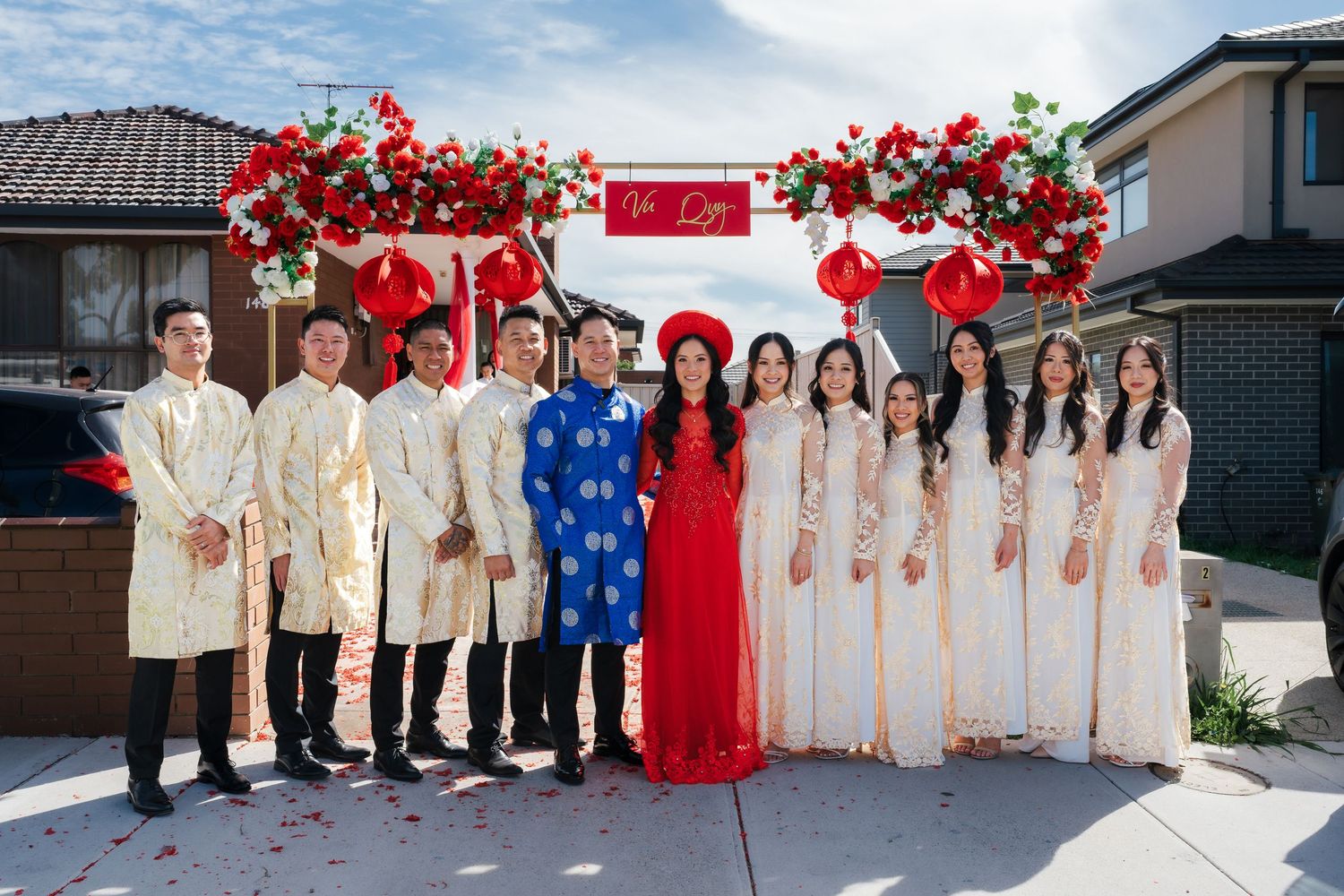 Wedding party in traditional Vietnamese ao dai attire posing under red floral arch outdoors.
