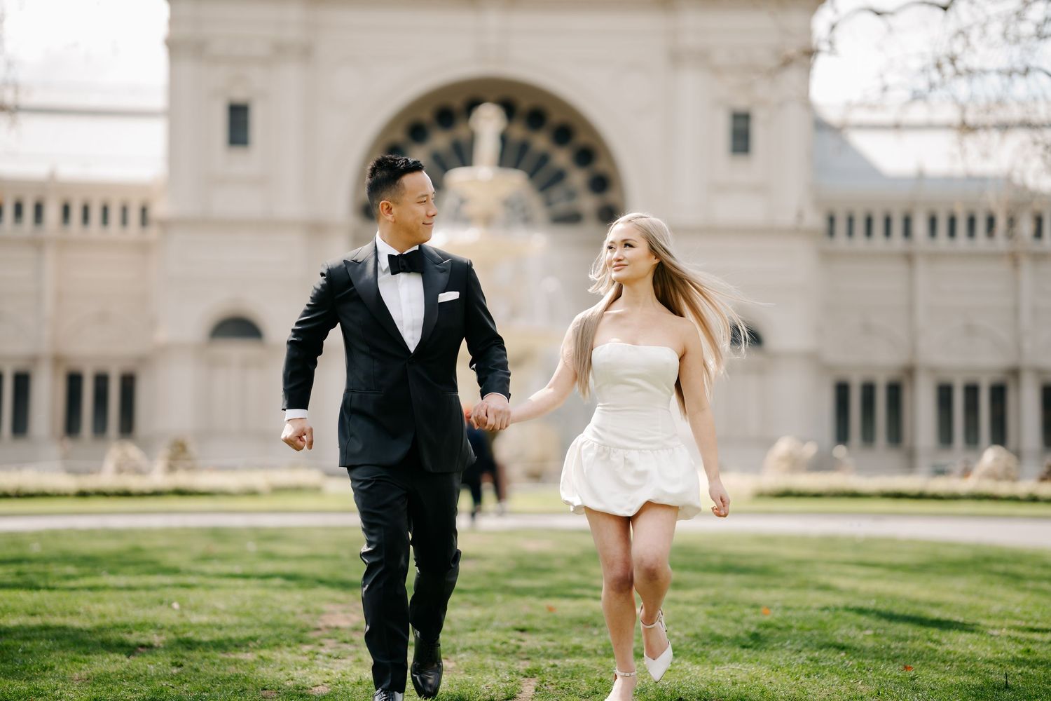 A joyful couple runs across a lawn with classic architecture in the background.