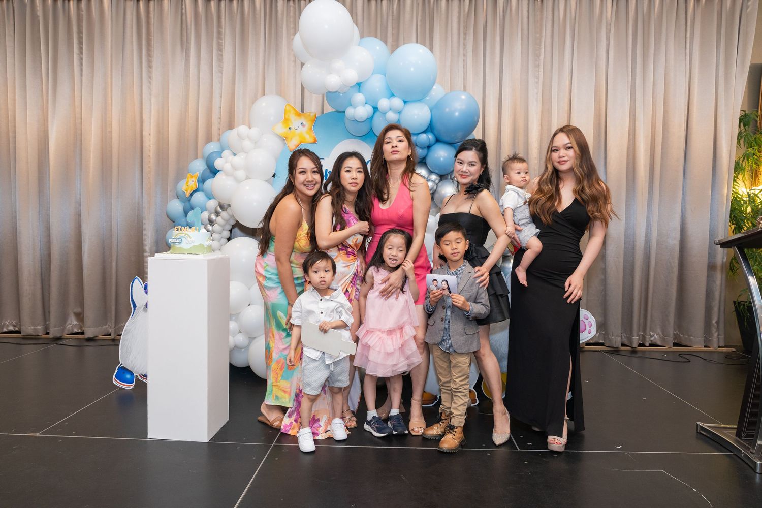 Group of women and children posing together at a festive baby shower with blue and white balloon arch decor.