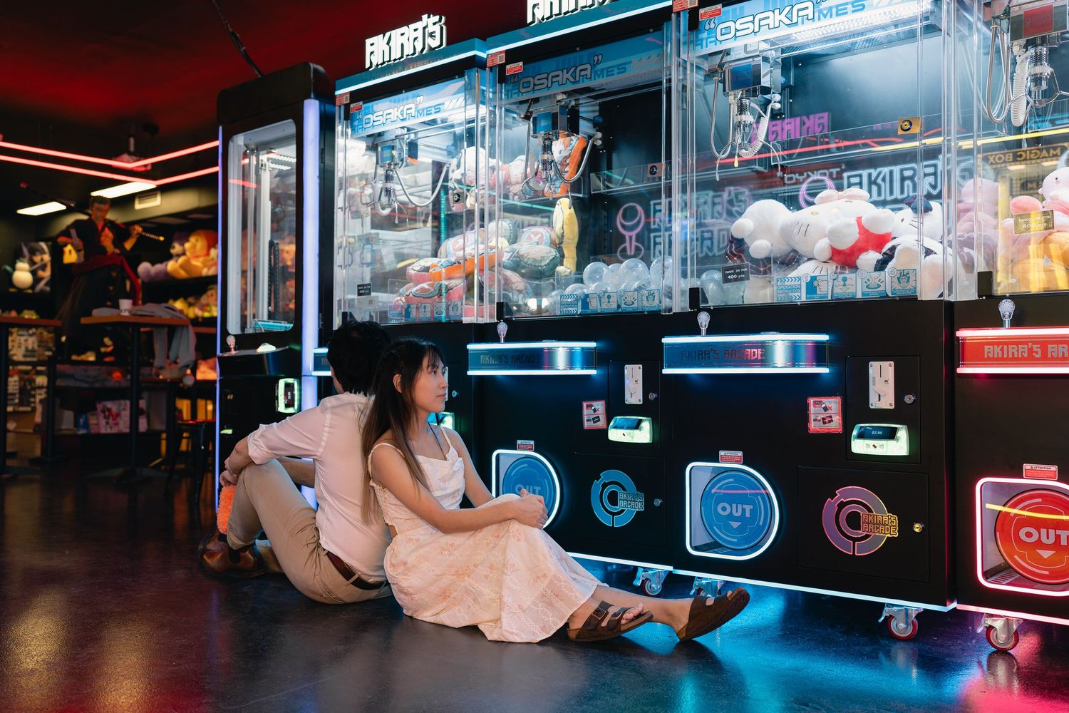 Two people sitting on the floor of a neon-lit arcade game center with claw machines.