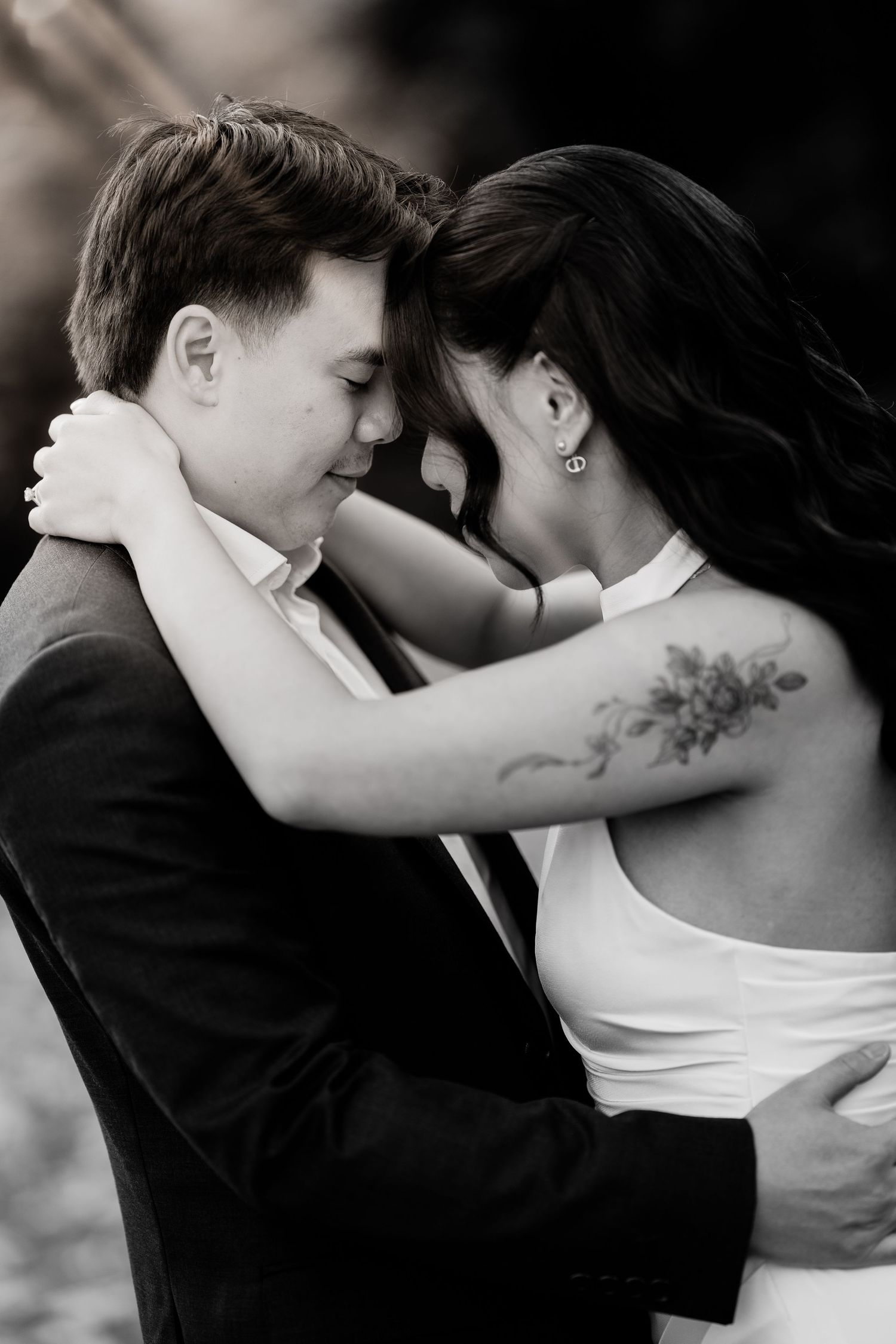 Black and white close-up of bride with floral tattoo embracing groom tenderly in outdoor setting.