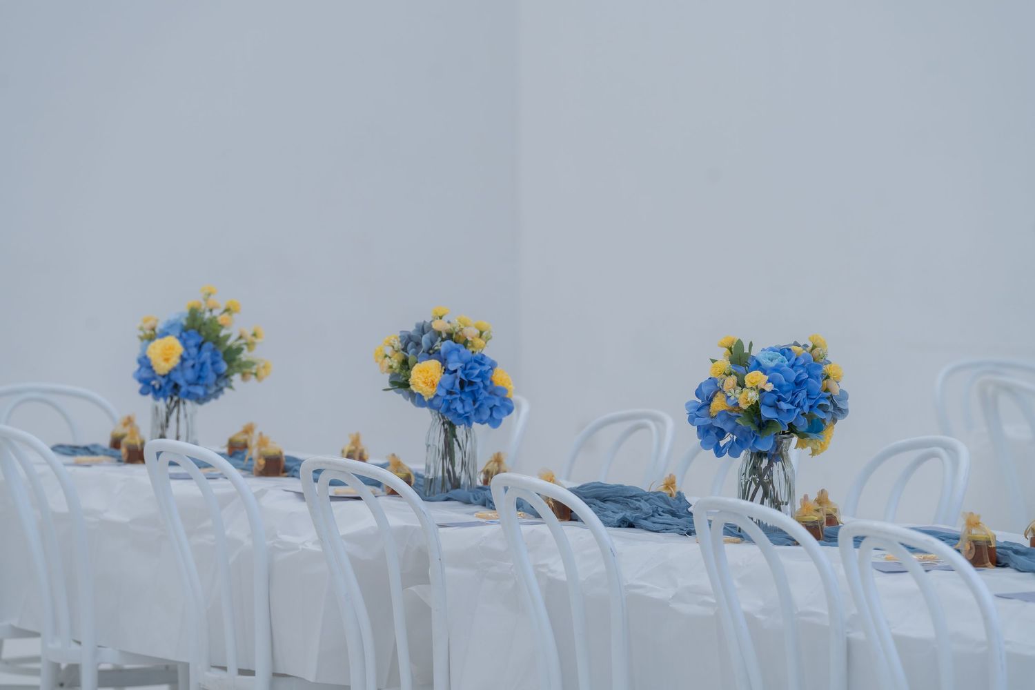 White chairs arranged at a table adorned with blue and yellow floral centerpieces in glass vases at an elegant event.