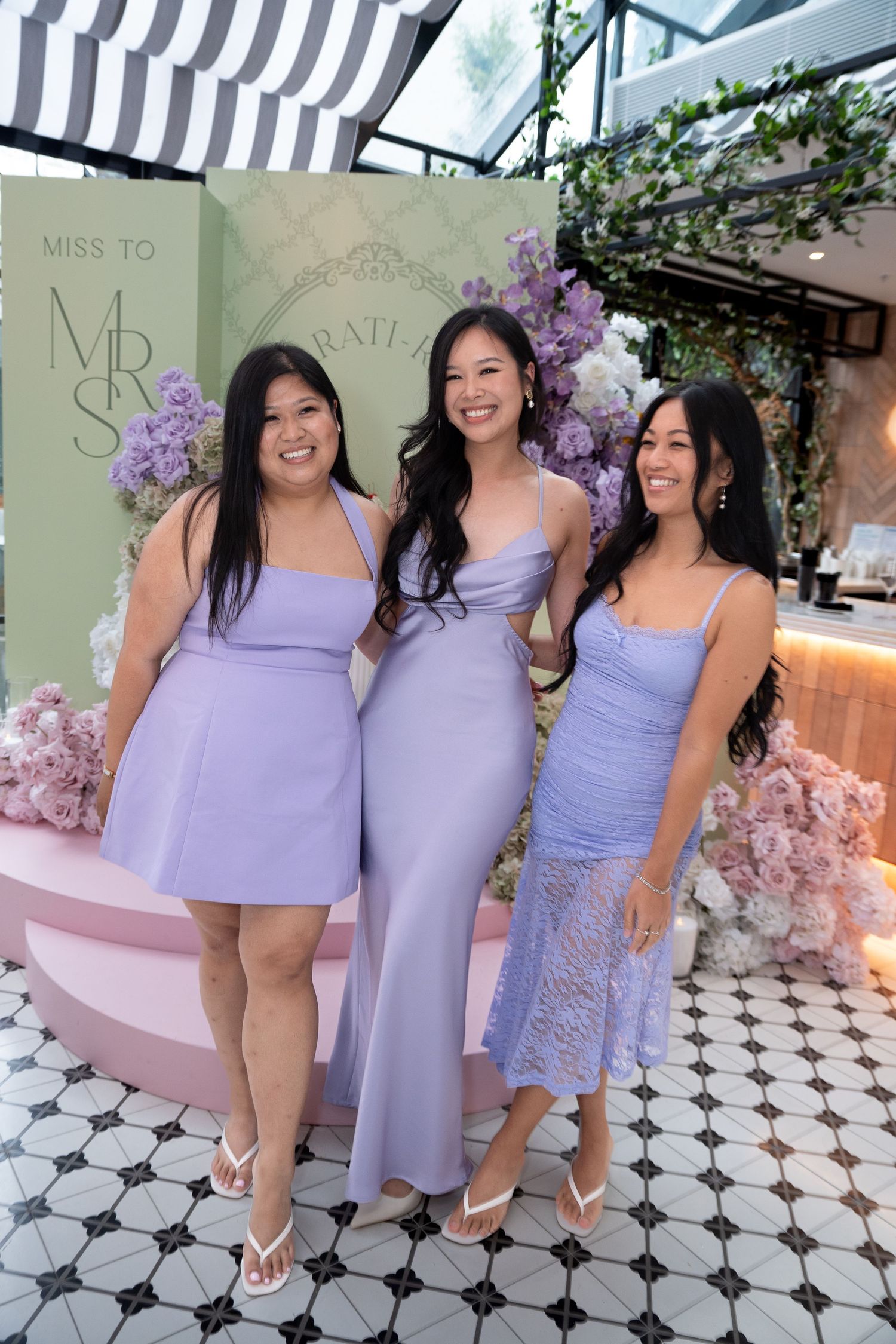 Three women in coordinating lavender dresses pose together at a bridal shower event with floral decor.