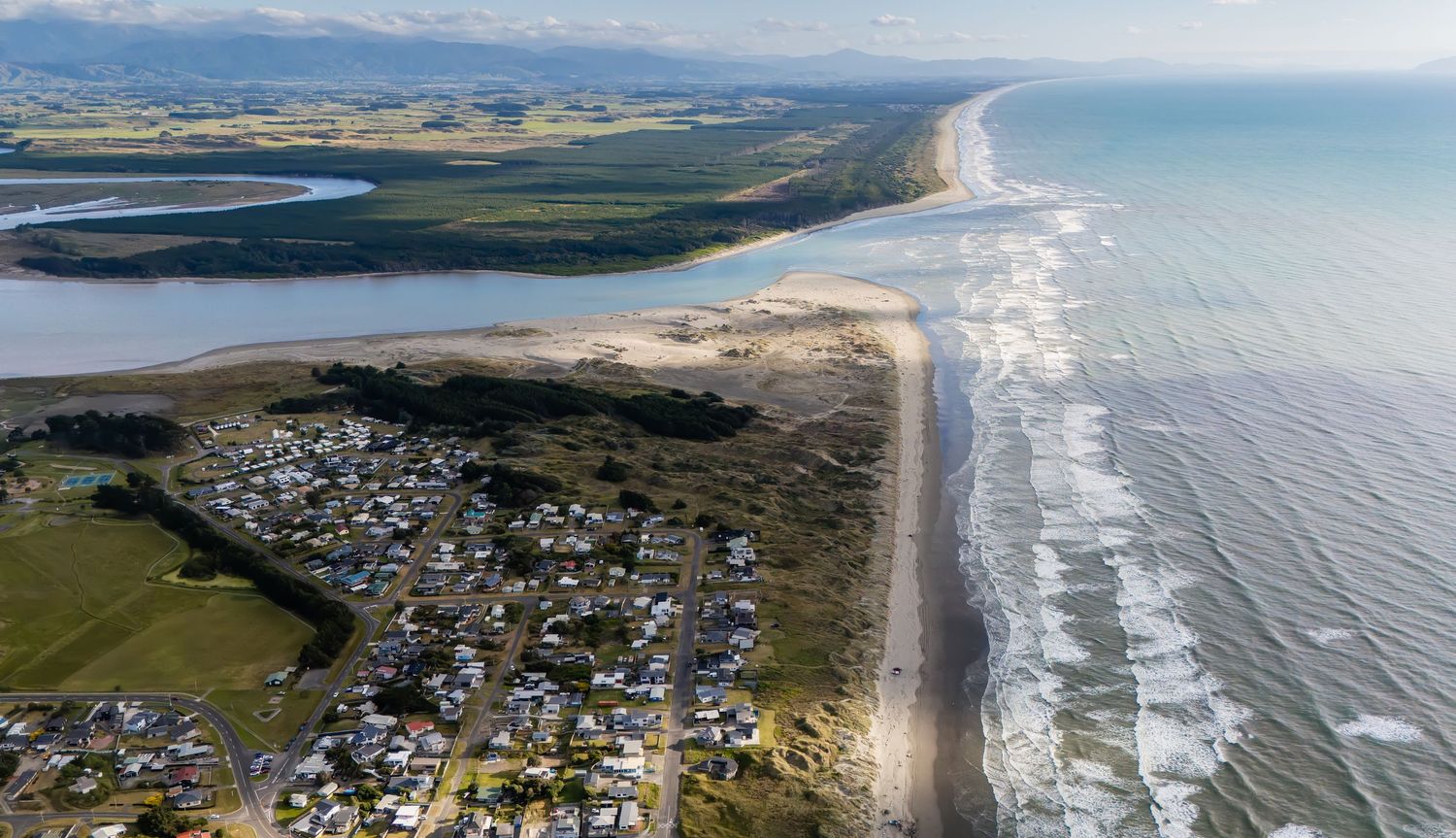 Aerial view of a New Zealand coastal settlement with long sandy beach and rolling ocean waves.