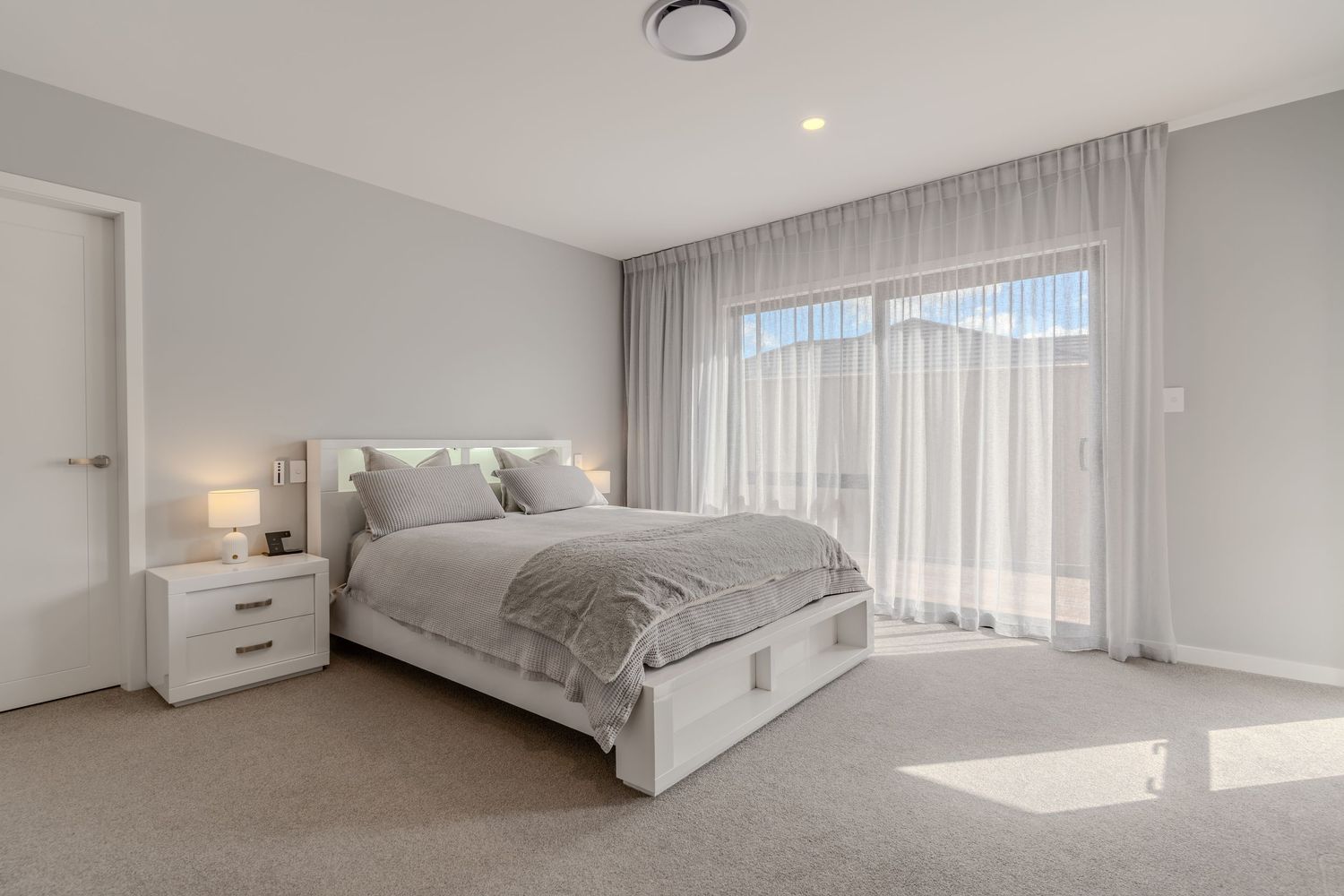 Bright modern bedroom with grey carpet, white storage bed, sheer curtains, and large window allowing natural light.