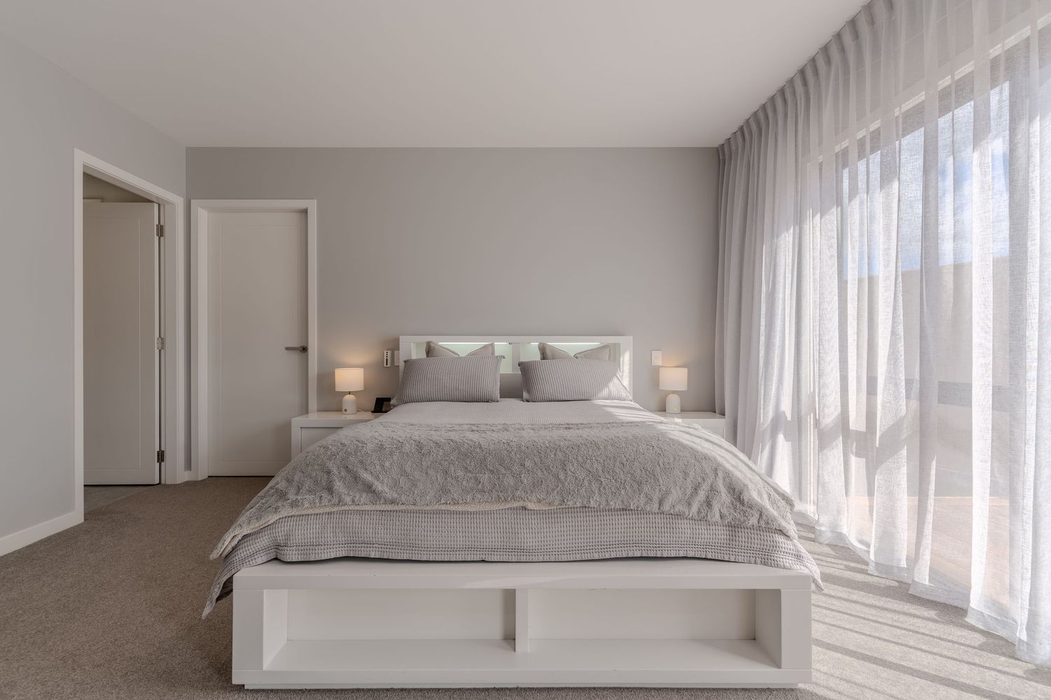 Bright minimalist bedroom with grey bedding, white furniture, sheer curtains, and neutral tones creating a serene atmosphere.