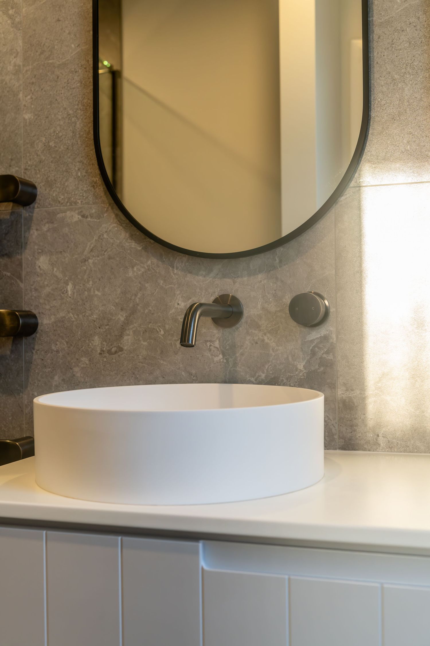 Modern bathroom vanity with round white vessel sink, wall-mounted faucet, gray marble tiles, and oval mirror.