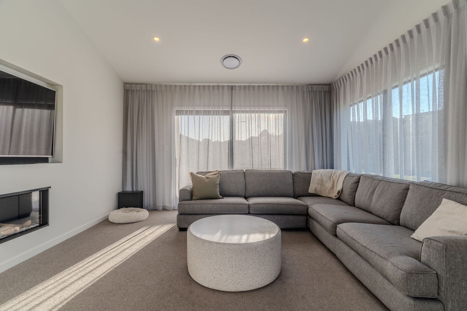 Modern living room with large grey sectional sofa, round ottoman, sheer curtains, and wall-mounted fireplace on white walls.