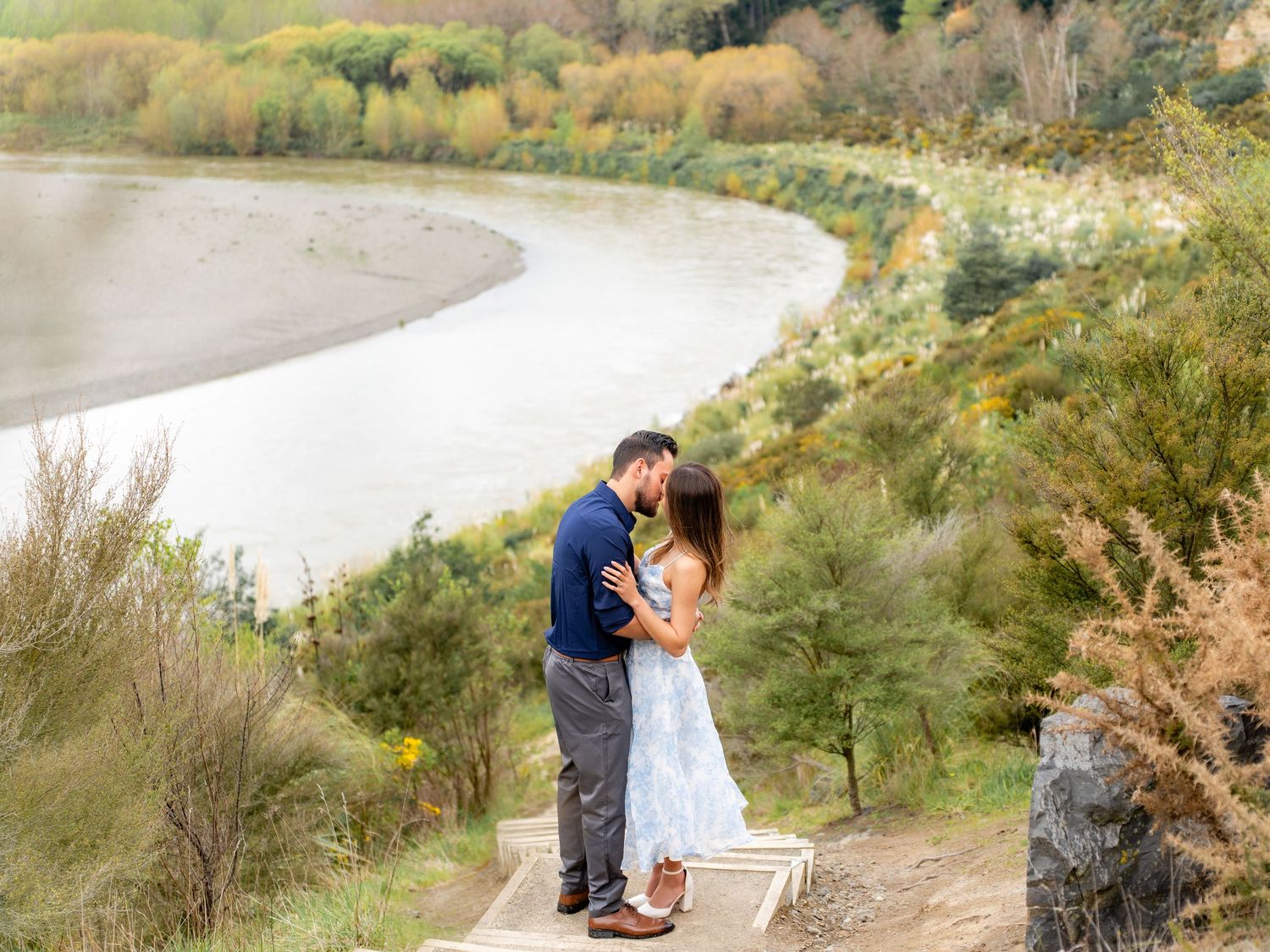 Couple sharing a kiss on overlook with winding river and lush green hills behind them.