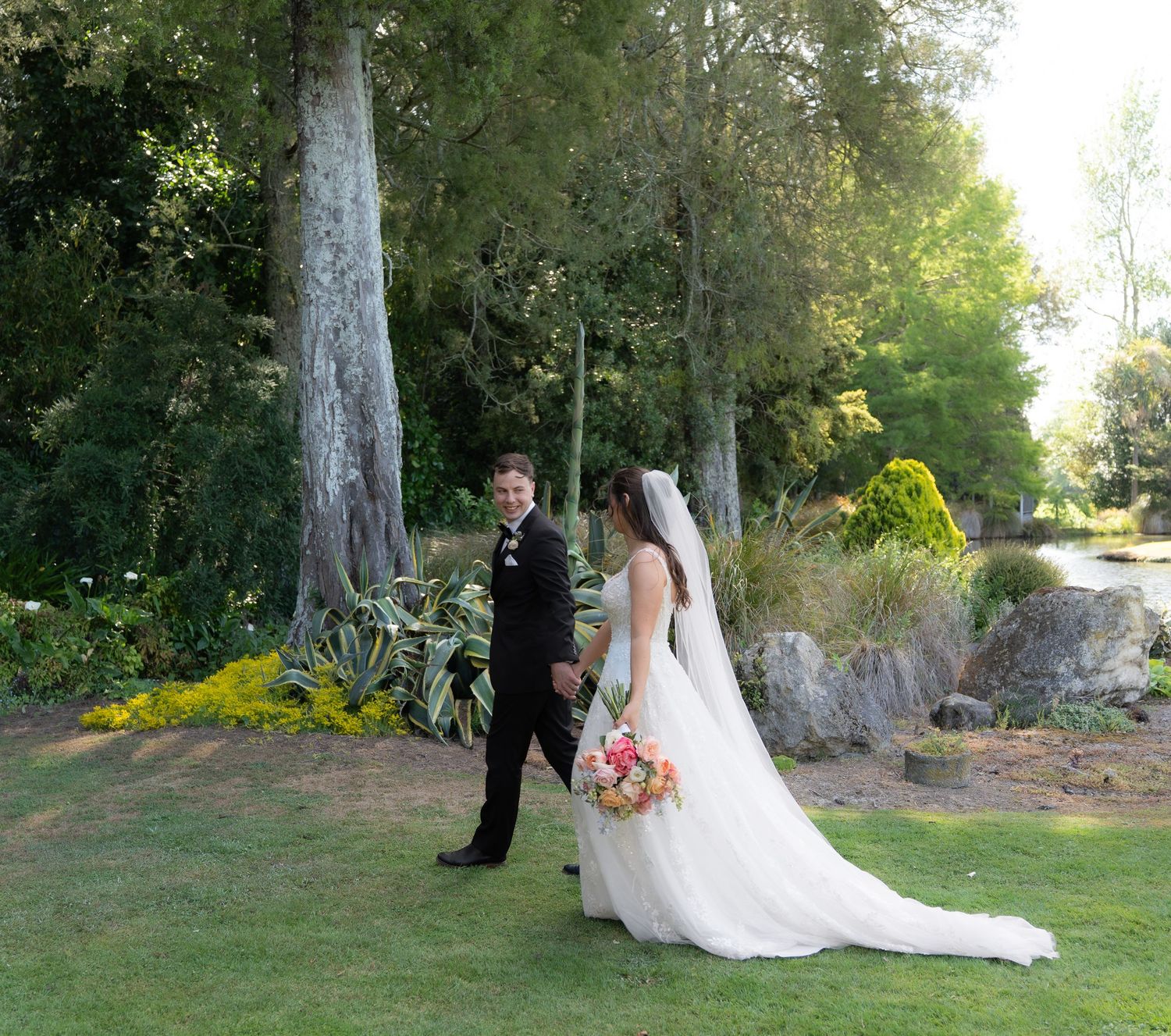 Bride and groom holding hands beside a scenic garden pond with tall trees.