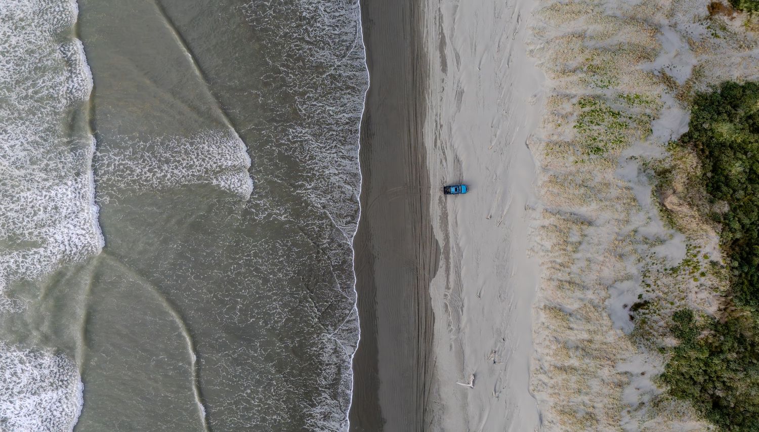 Aerial top-down view of a blue vehicle driving along a remote sandy beach beside ocean waves at foxton beach.